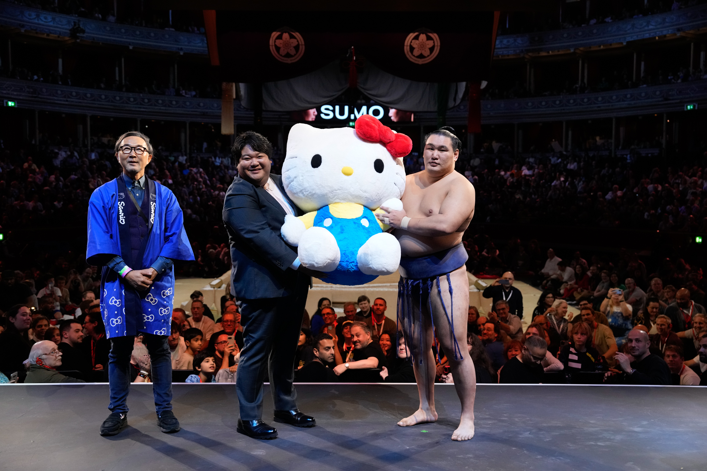 Three people posing with large white and blue Hello Kitty mascot in sumo wrestling arena with crowd in background.