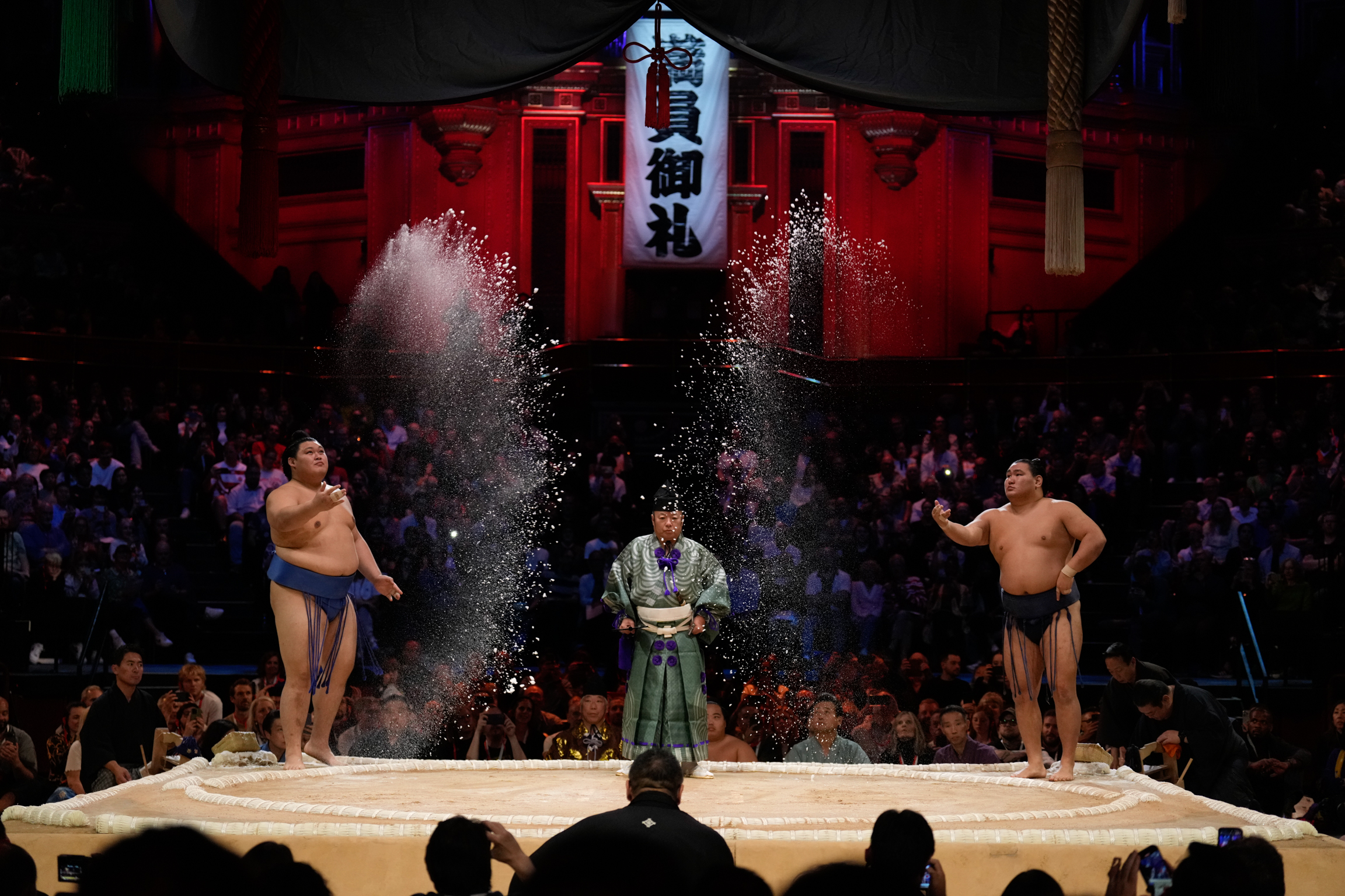 Two sumo wrestlers throw salt whilst referee in green robes stands between them on ring. Red shrine backdrop, crowd watching.