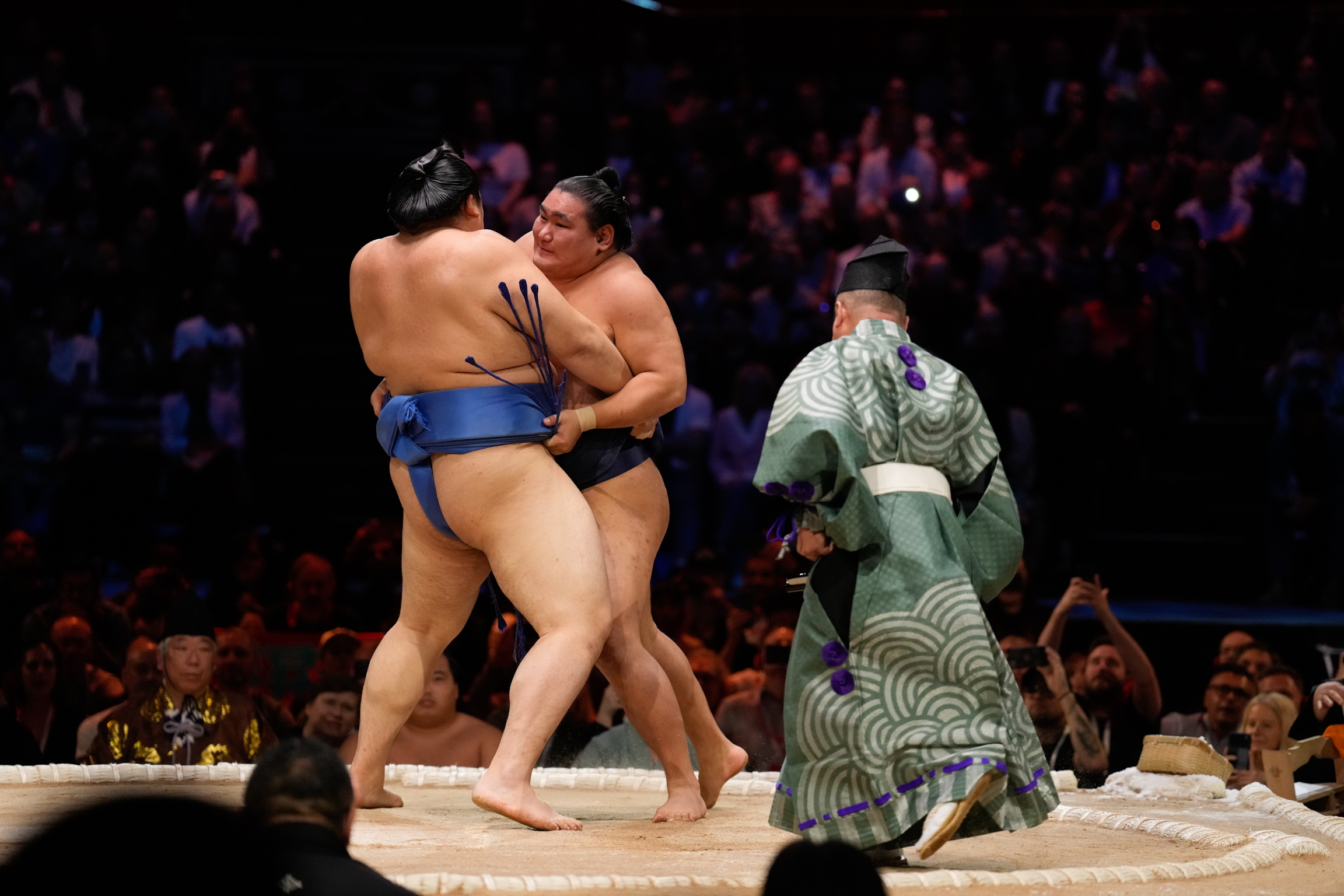 Two sumo wrestlers grappling on sandy ring whilst referee in green patterned robe watches, crowd visible in dark background.