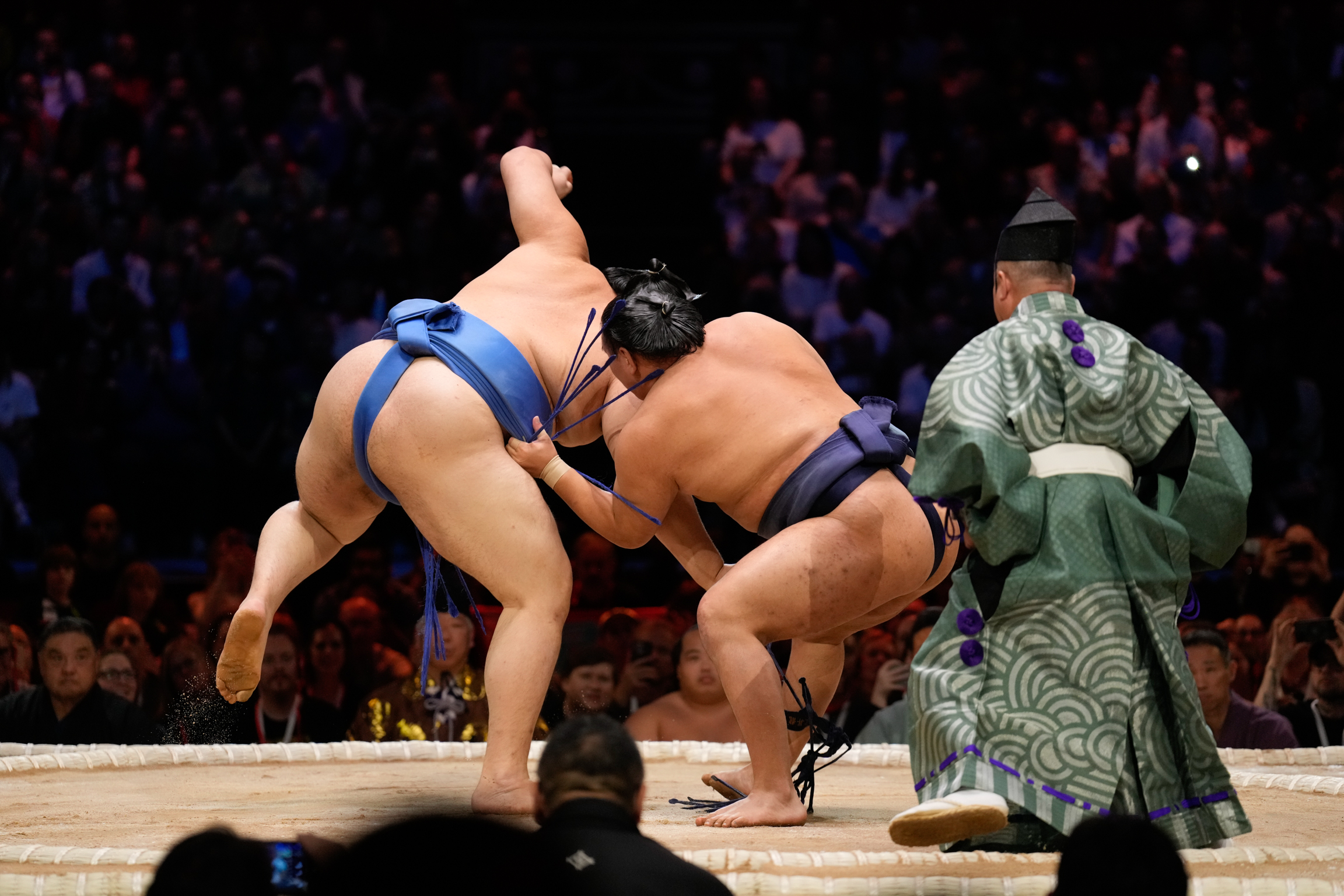 Two sumo wrestlers grappling on sandy ring whilst referee in green patterned kimono observes, with crowd in background.