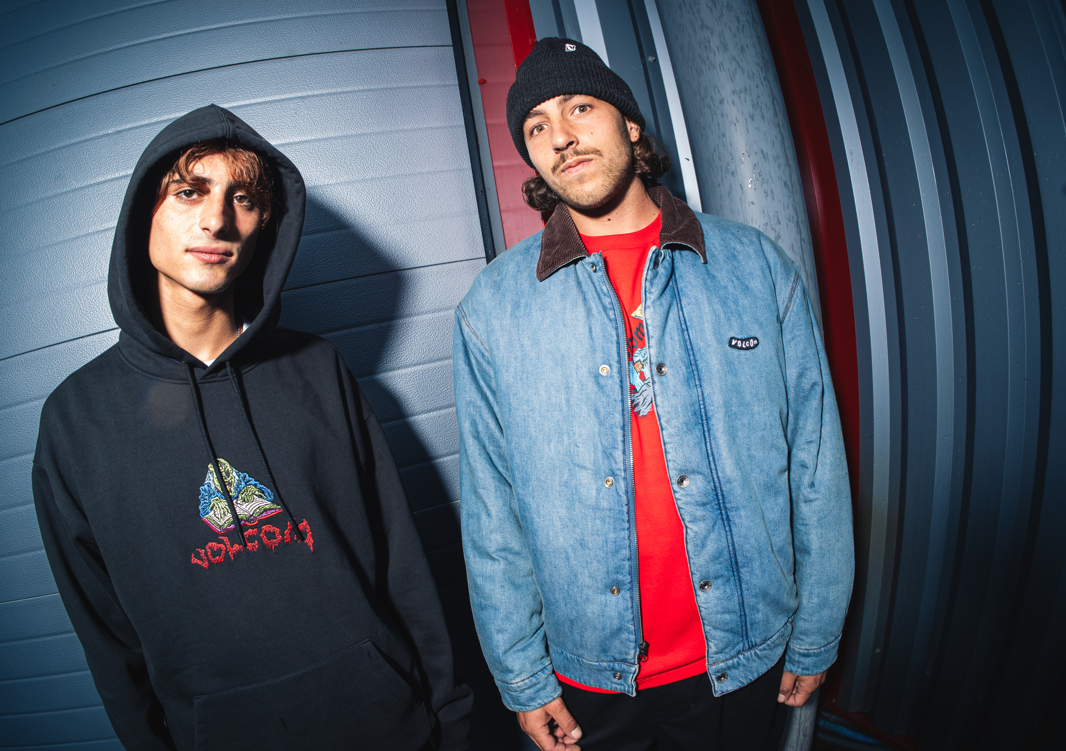 Two men in casual wear standing against curved metallic wall. Left wears black hoodie, right wears denim jacket over red shirt and beanie.
