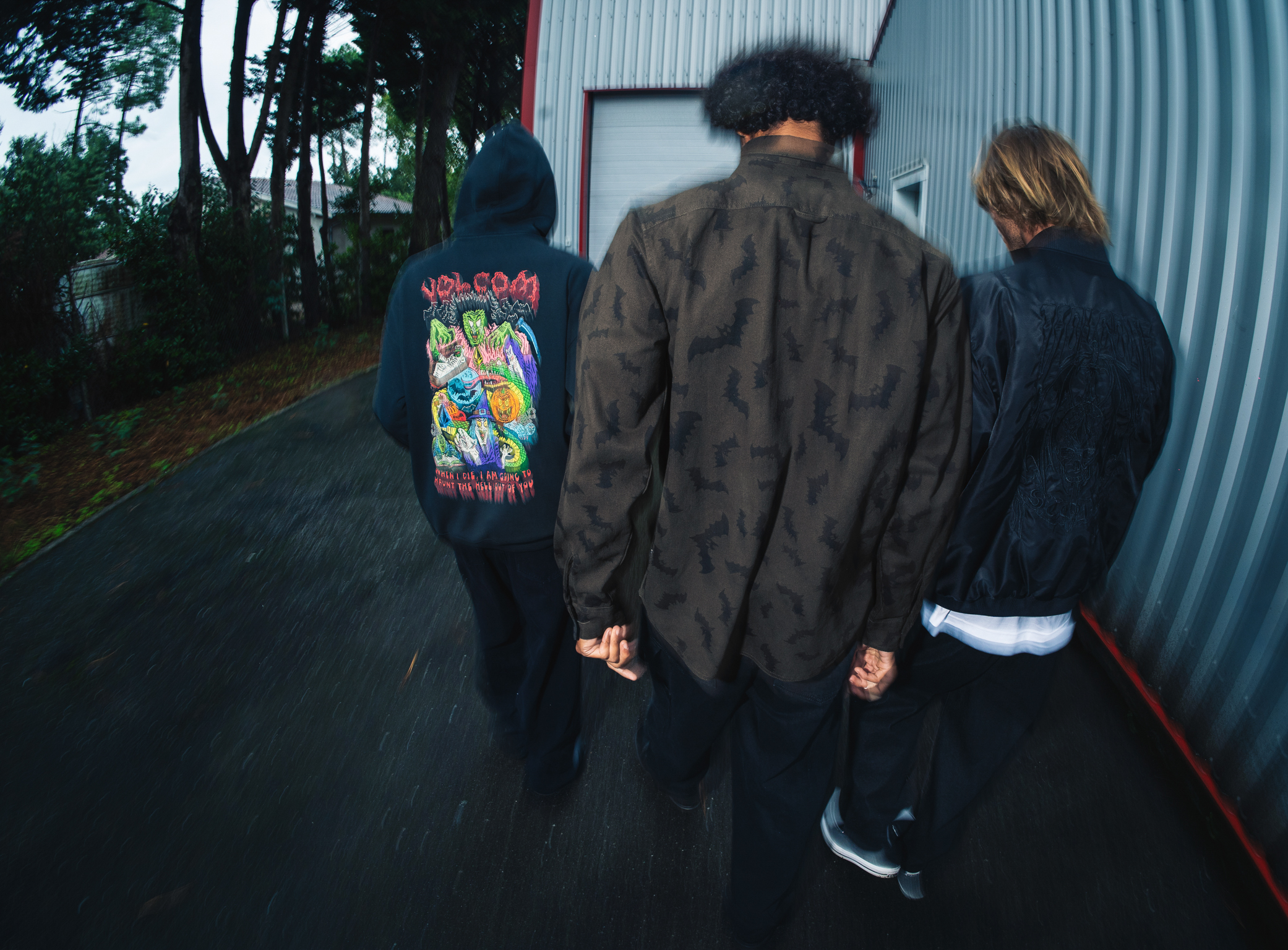 Three people walking away from camera towards corrugated metal building. Person on left wears colourful graphic t-shirt, centre person in camouflage jacket.