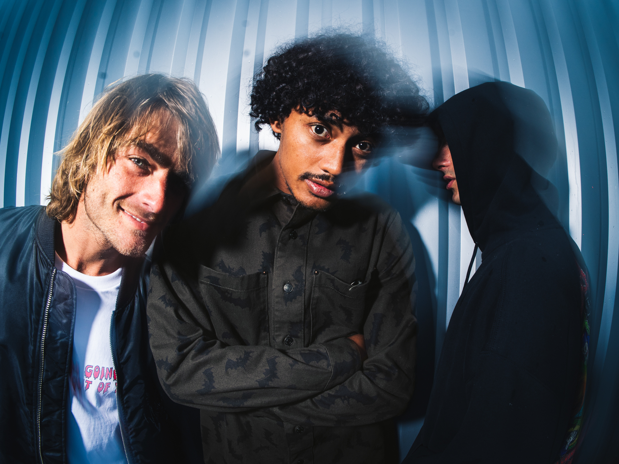 Three young men posing together against blue striped backdrop, with blonde, curly dark, and obscured hair styles in casual clothing.