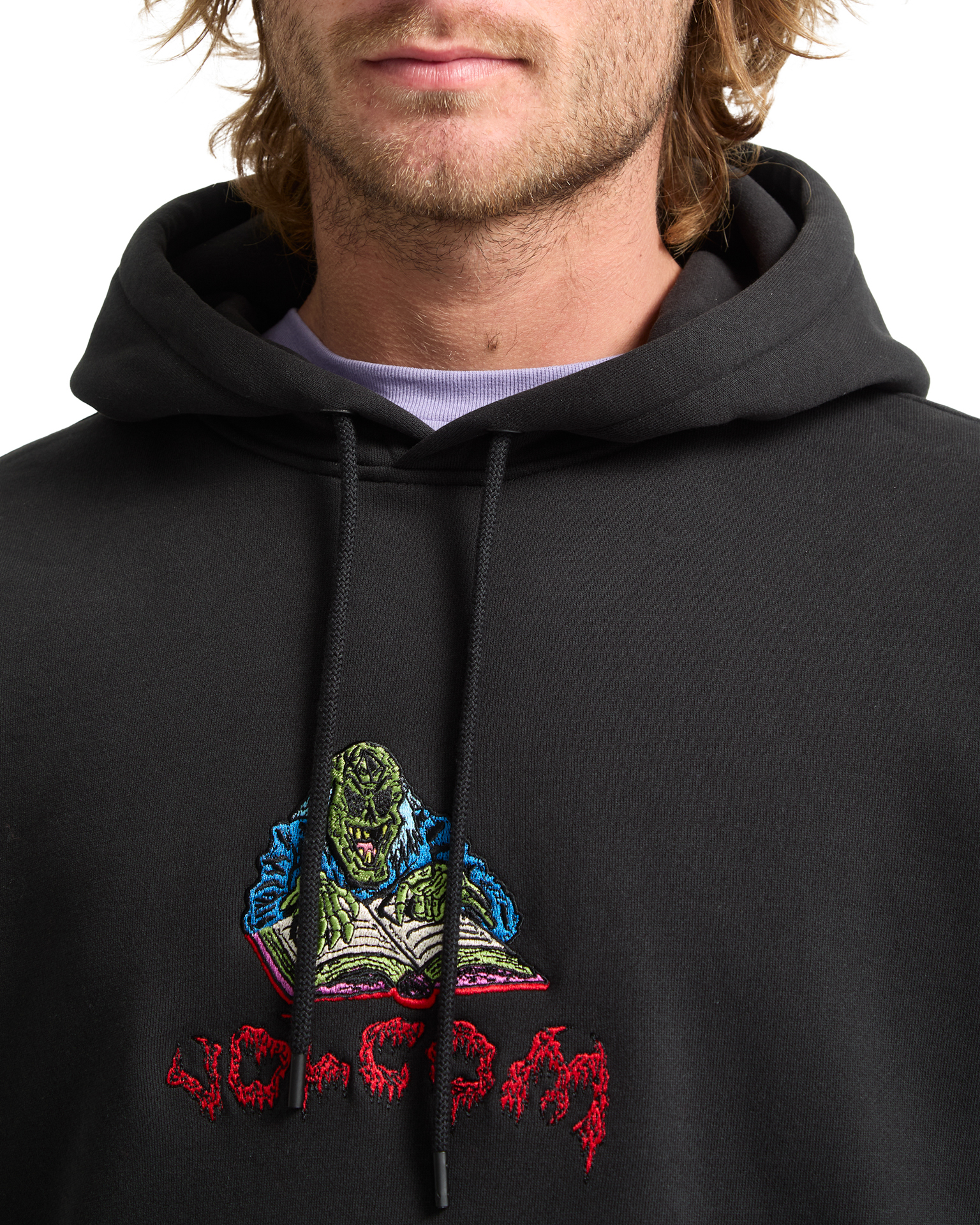 Man wearing black hoodie with embroidered monster character in green and blue colours above red gothic text.