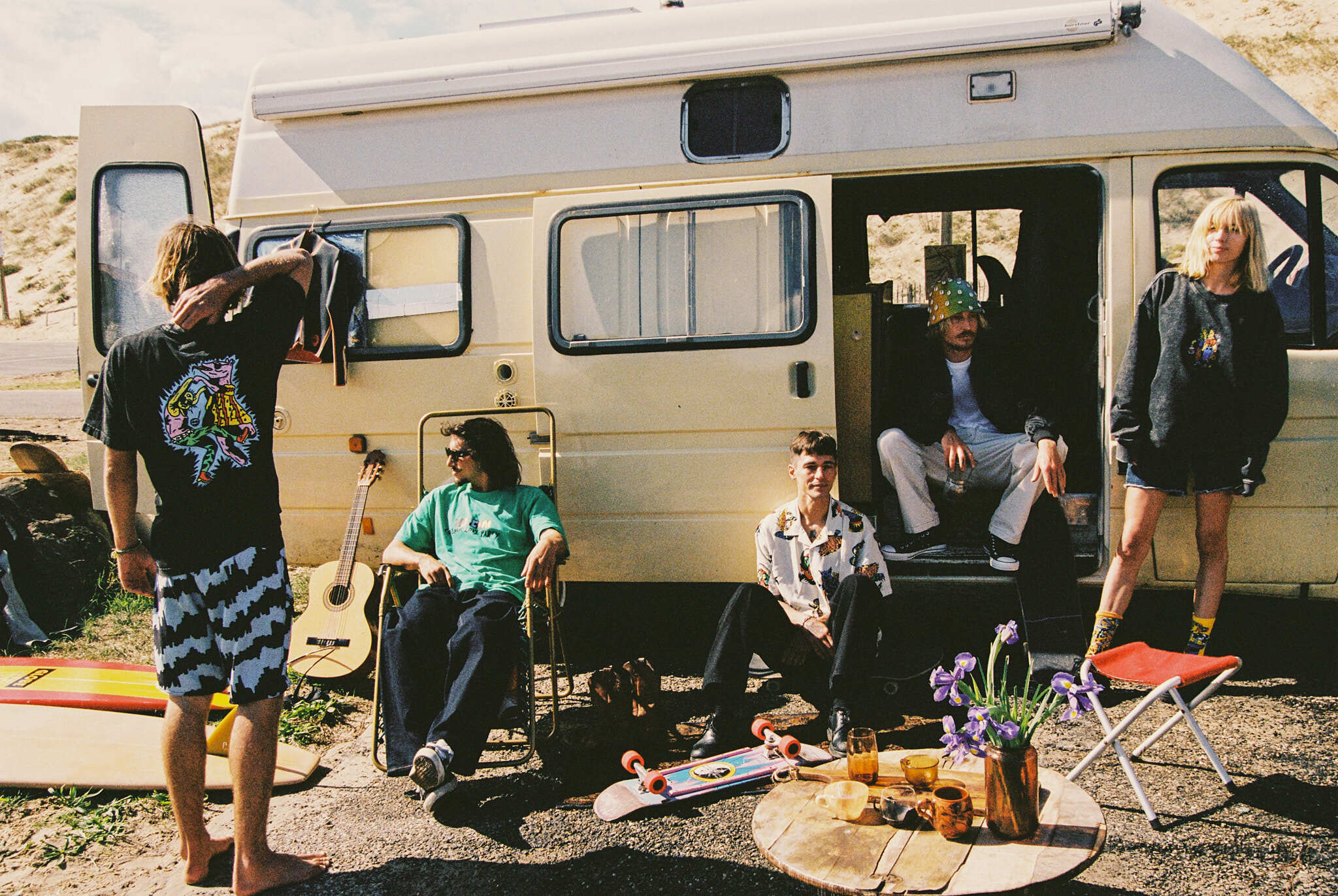 Image shows a group of people in and around a beige campervan in a desert setting. Four people are visible - two standing outside the van and two seated inside. Several camping items like chairs and guitar are visible on the ground in front of the van.