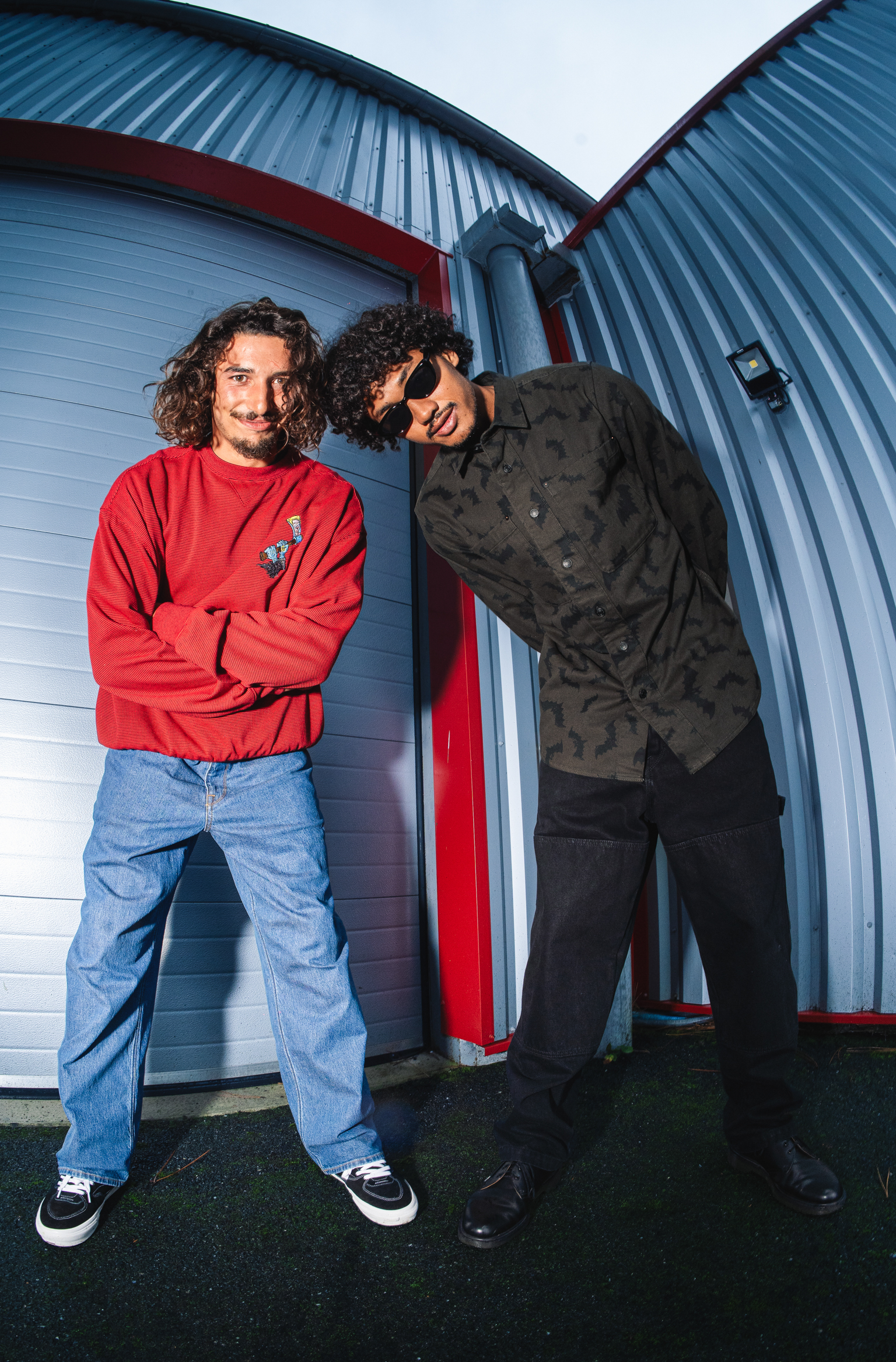 Two men with curly hair standing against corrugated metal building with red trim. One wears red jumper and jeans, other in dark shirt.