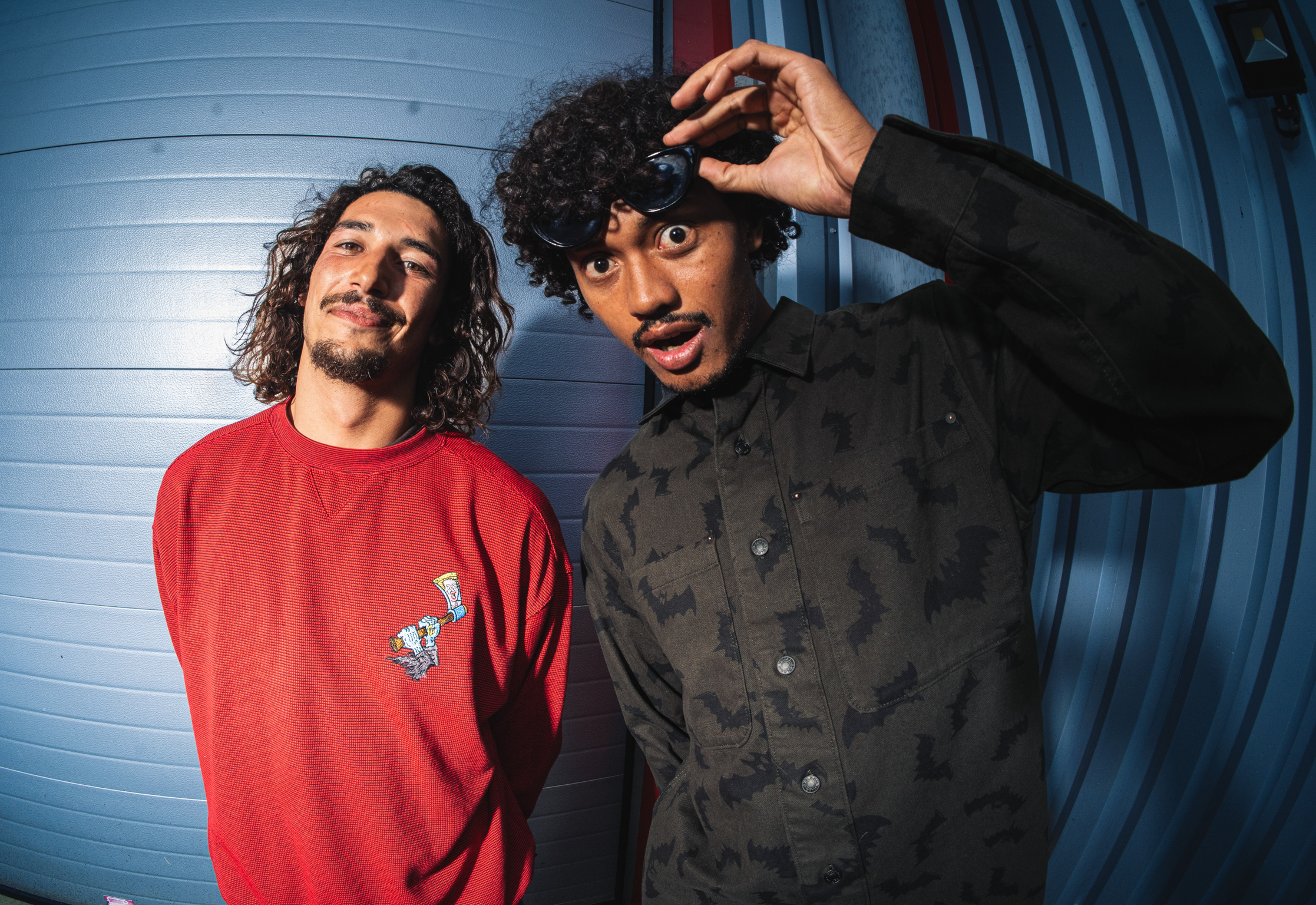 Two men with curly hair against blue wooden wall. Left wears red jumper, right wears black shirt with hand raised to head.