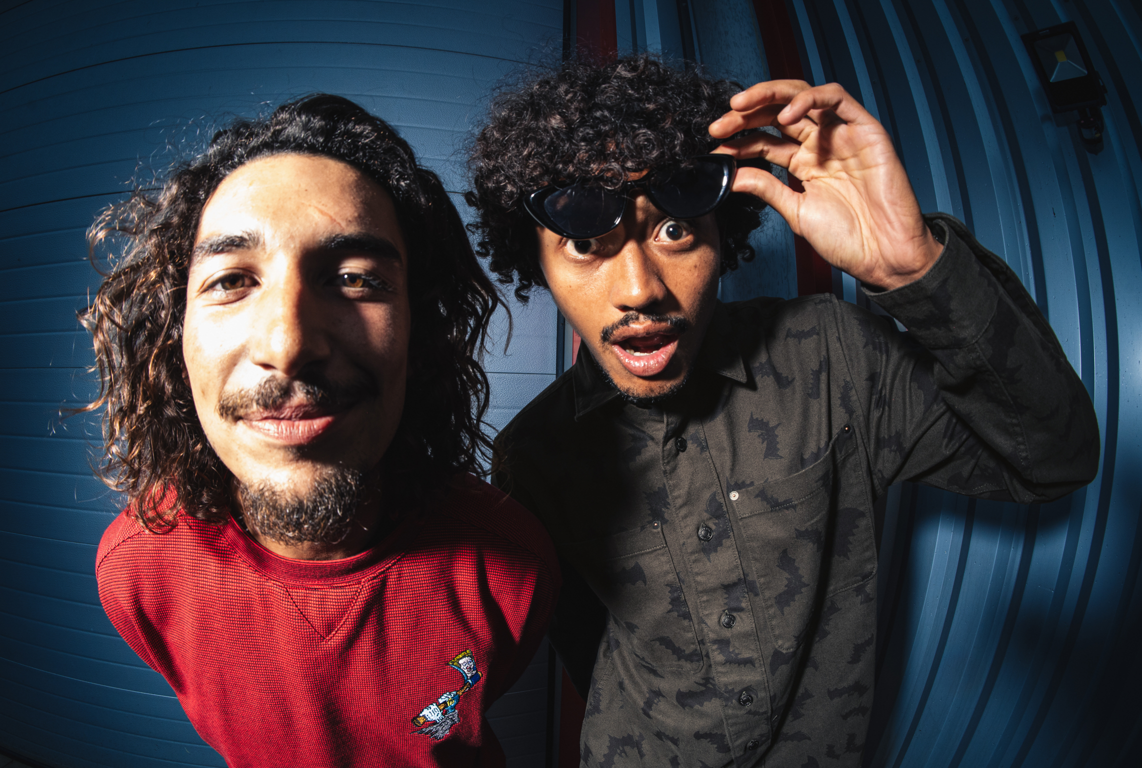 Two men with curly hair, one in red jumper, one in black shirt wearing sunglasses and adjusting them with his hand.