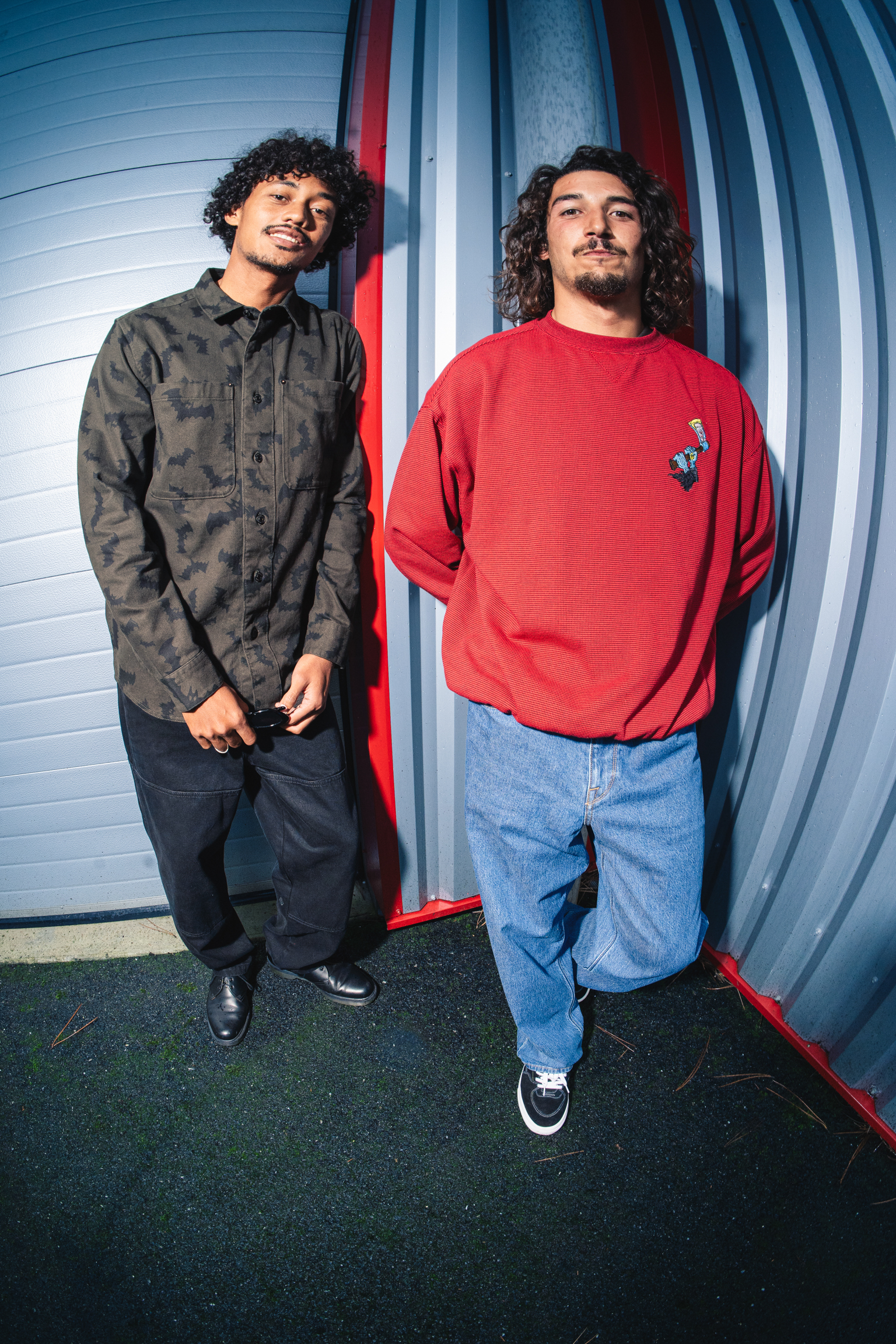 Two men standing against curved metal wall with red and blue stripes. Left wears camouflage shirt, right wears red jumper and jeans.