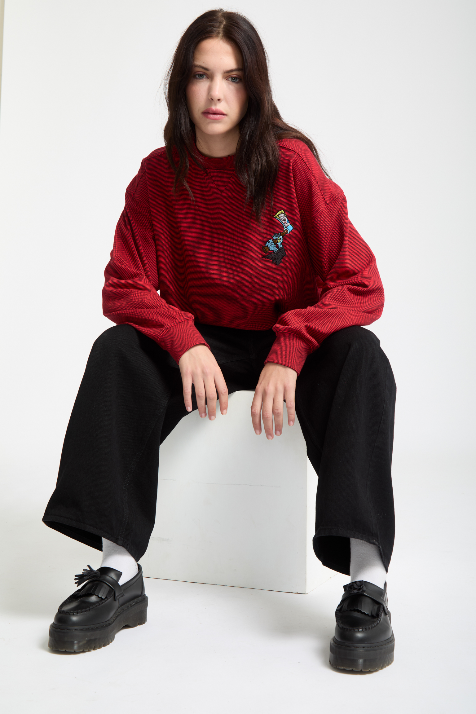 Woman with long dark hair sitting on white block wearing red jumper with small graphic, black trousers, and black shoes against white background.