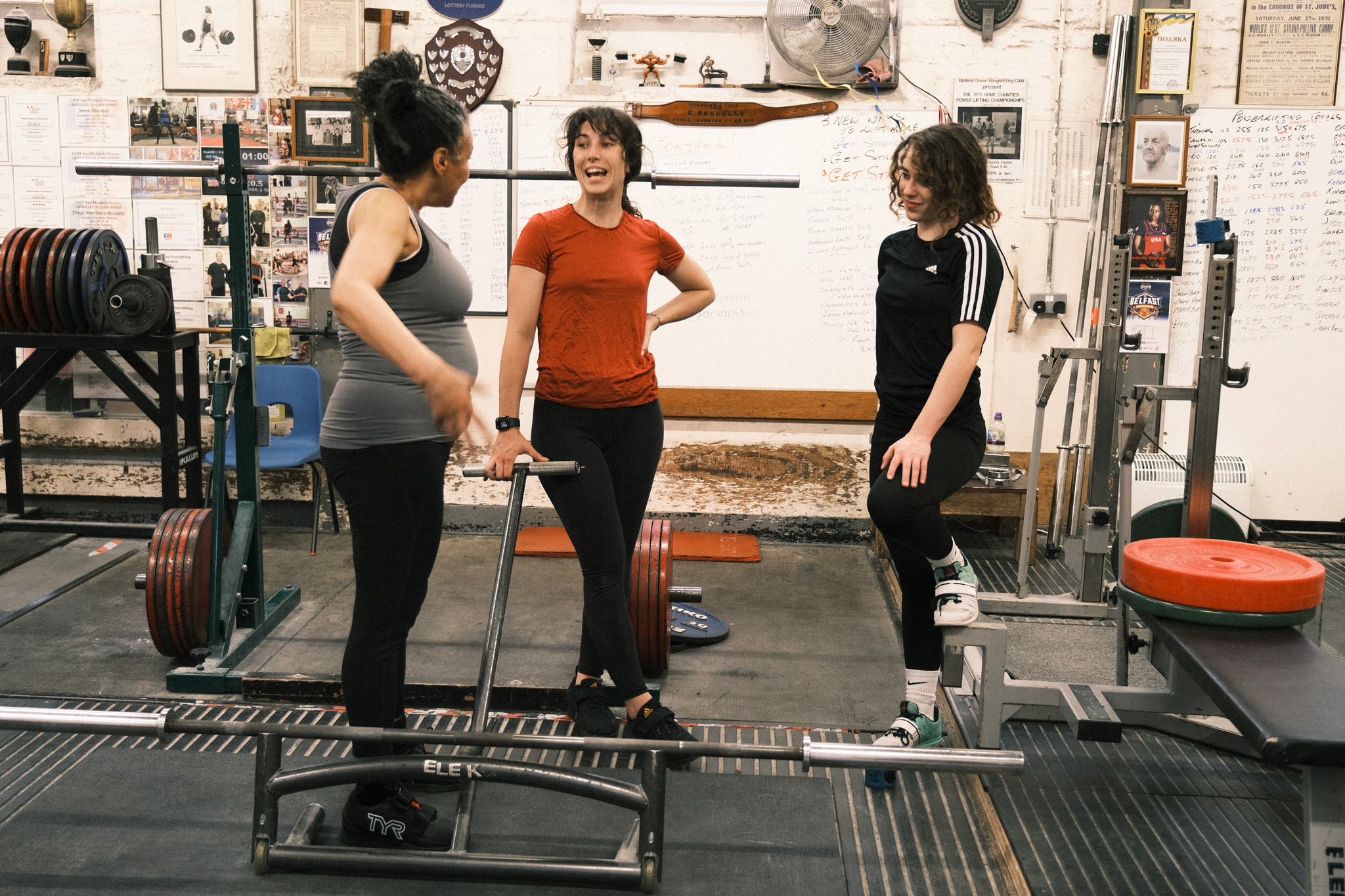 Three women in a gym with weightlifting equipment, one in grey vest, one in red top, one in black Adidas tracksuit.