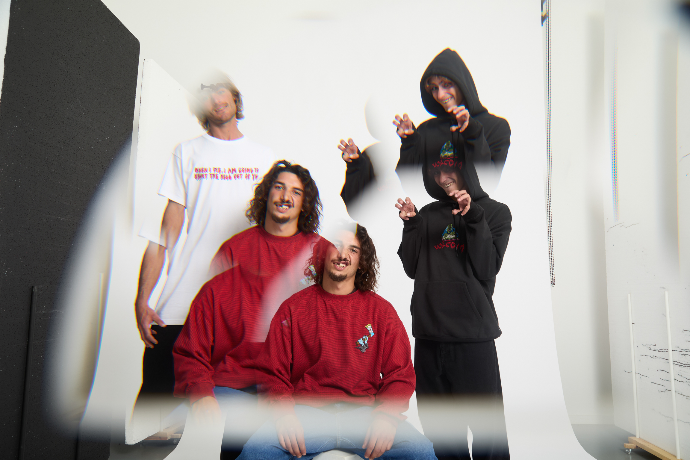 Four people in casual clothing: one in white t-shirt, two in red jumpers, one in black hoodie. Indoor setting with white walls.