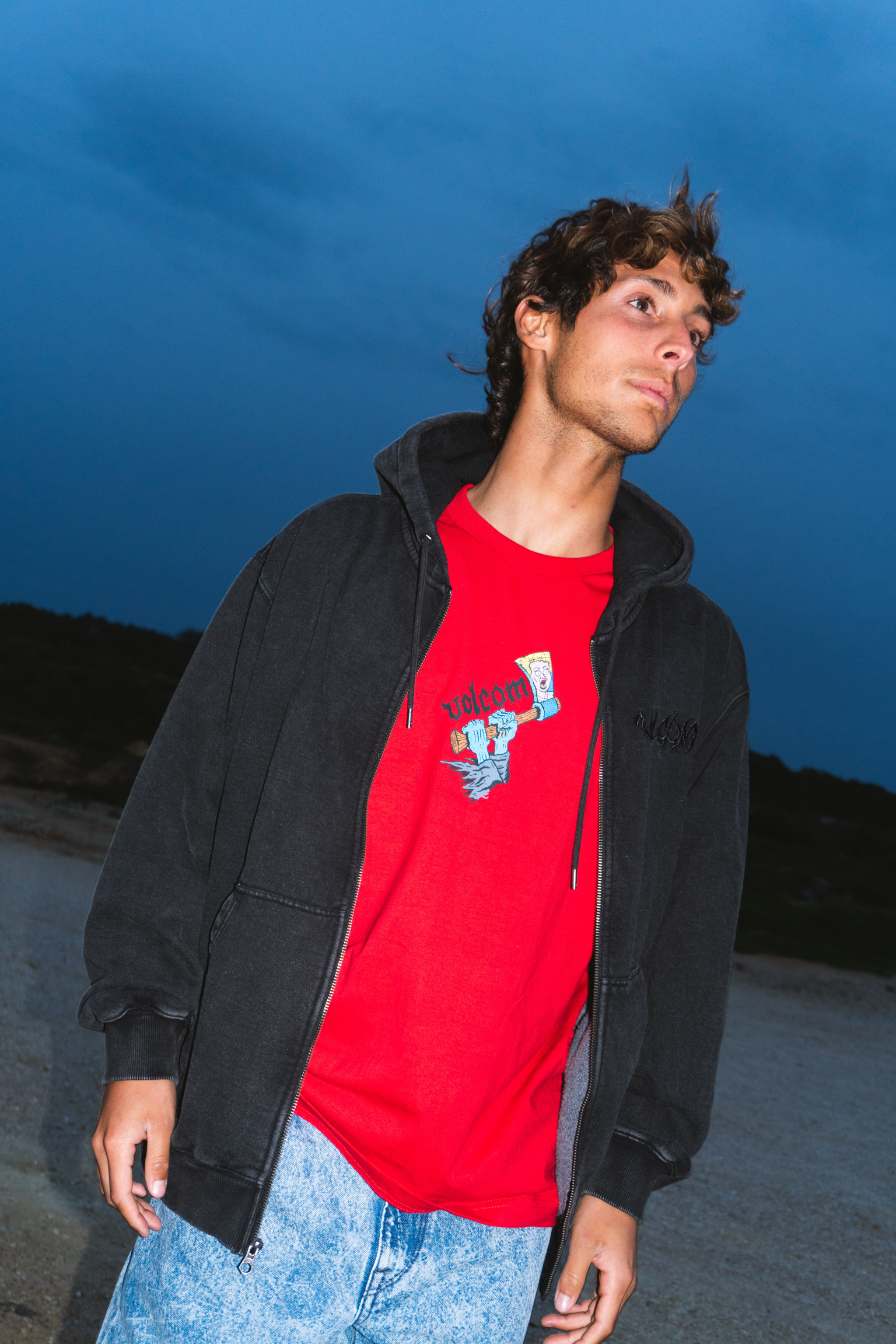 Young man in black hoodie and red graphic t-shirt stands against blue evening sky, looking upward with tousled dark hair.