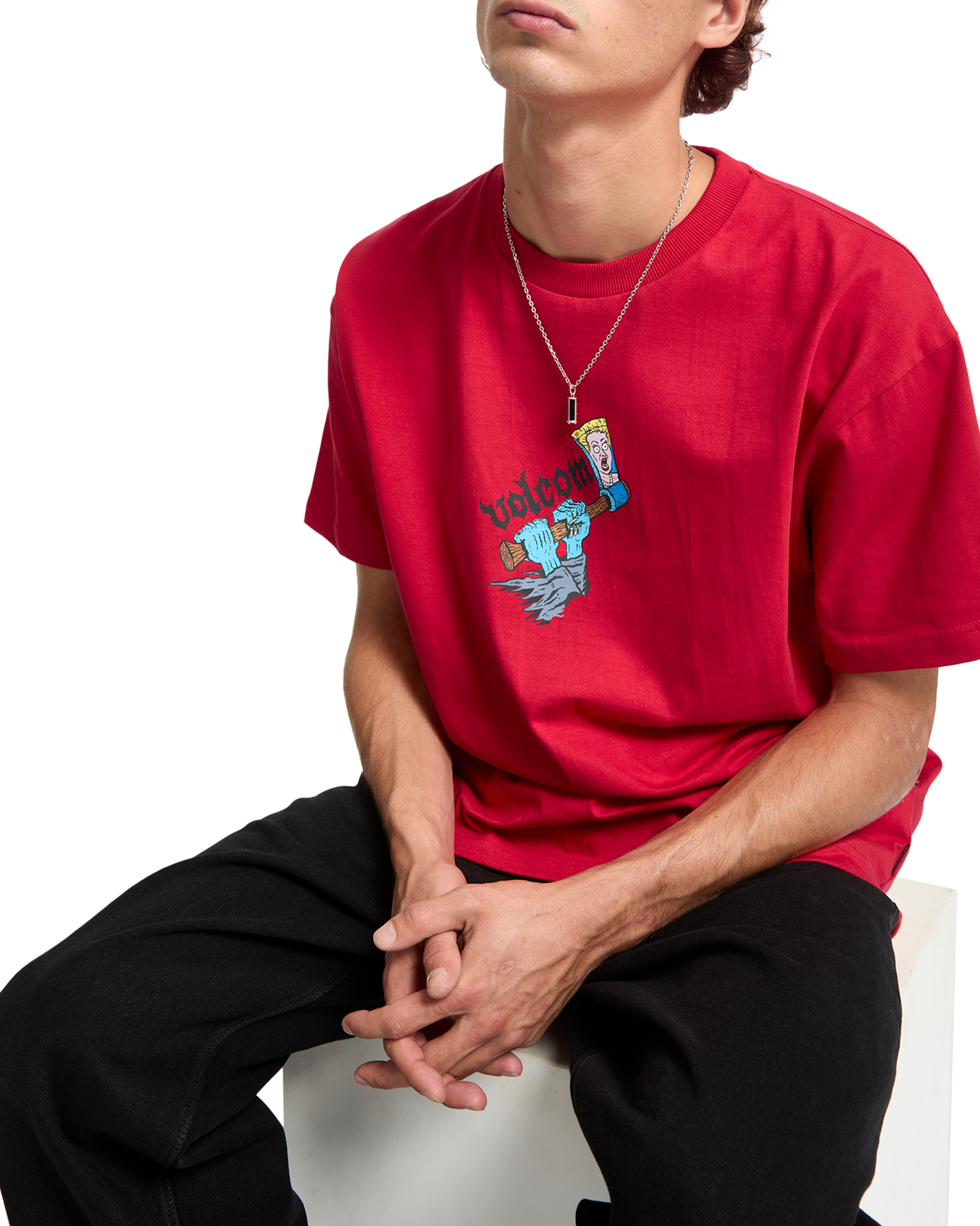 Person wearing red t-shirt with cartoon graphic and gold necklace, seated in black trousers against white background.