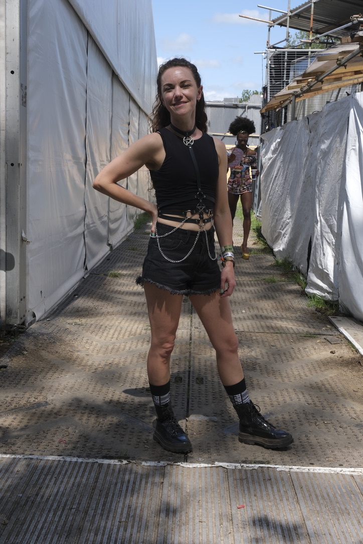 Woman in black crop top and denim skirt posing on metal walkway between white fabric barriers at outdoor event.