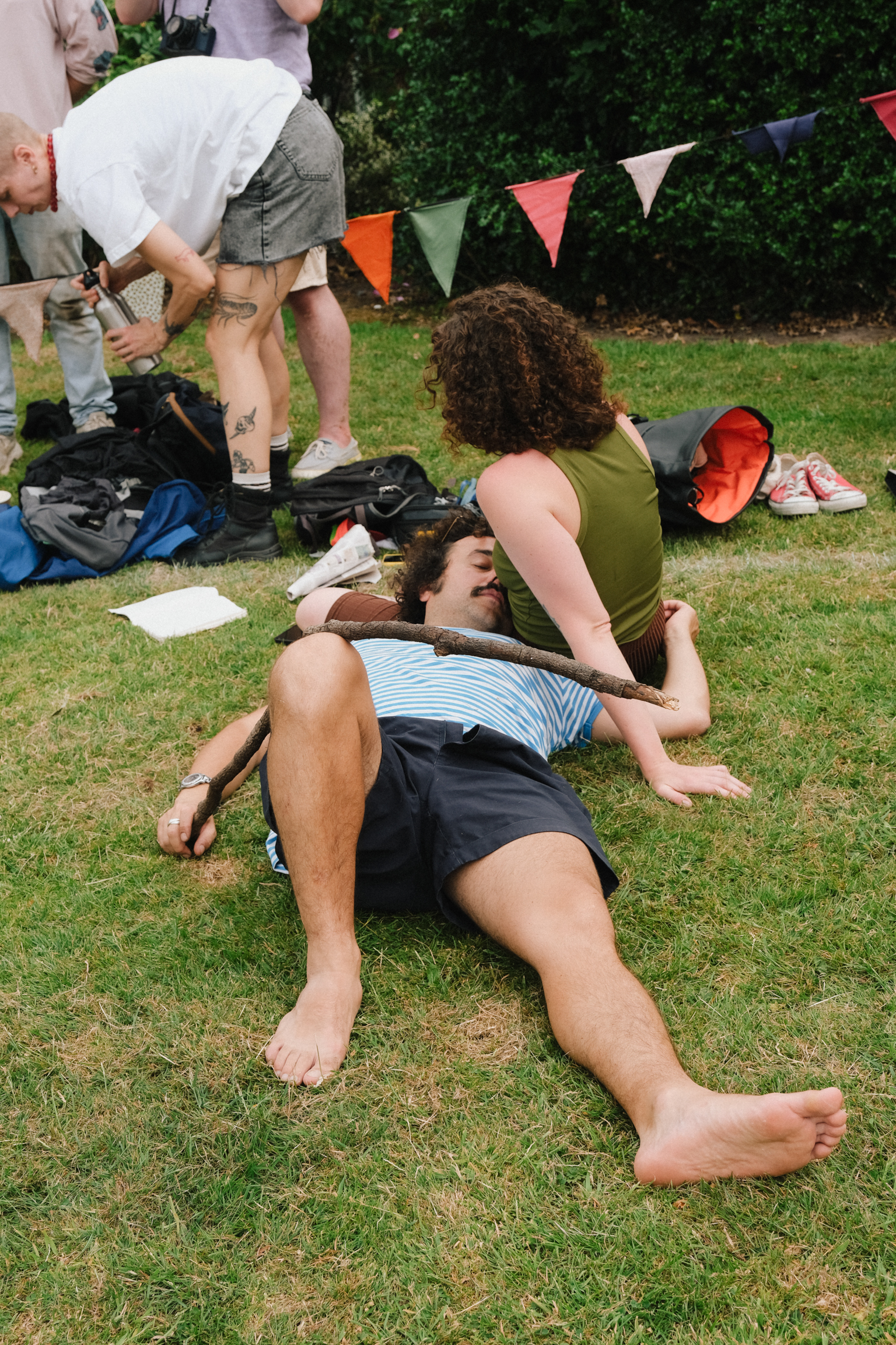 People relaxing on grass at outdoor event with colourful bunting. Woman in green top sits beside man lying down in striped shirt.