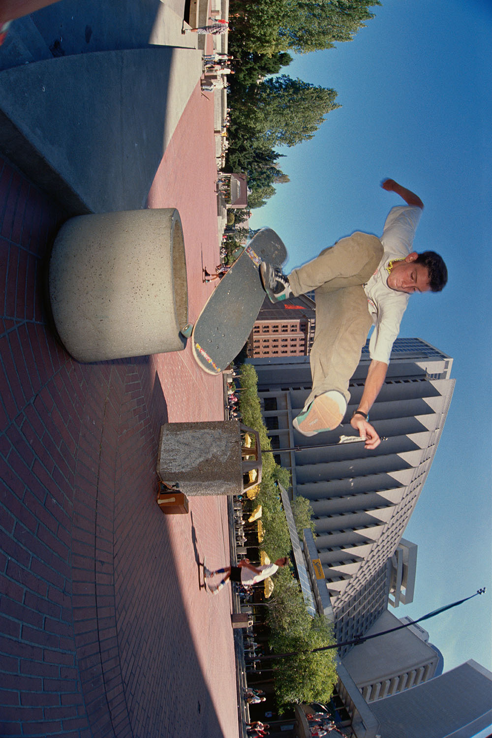 Man in white shirt and beige trousers standing on promenade beside large grey sculpture, with buildings and blue sky behind.