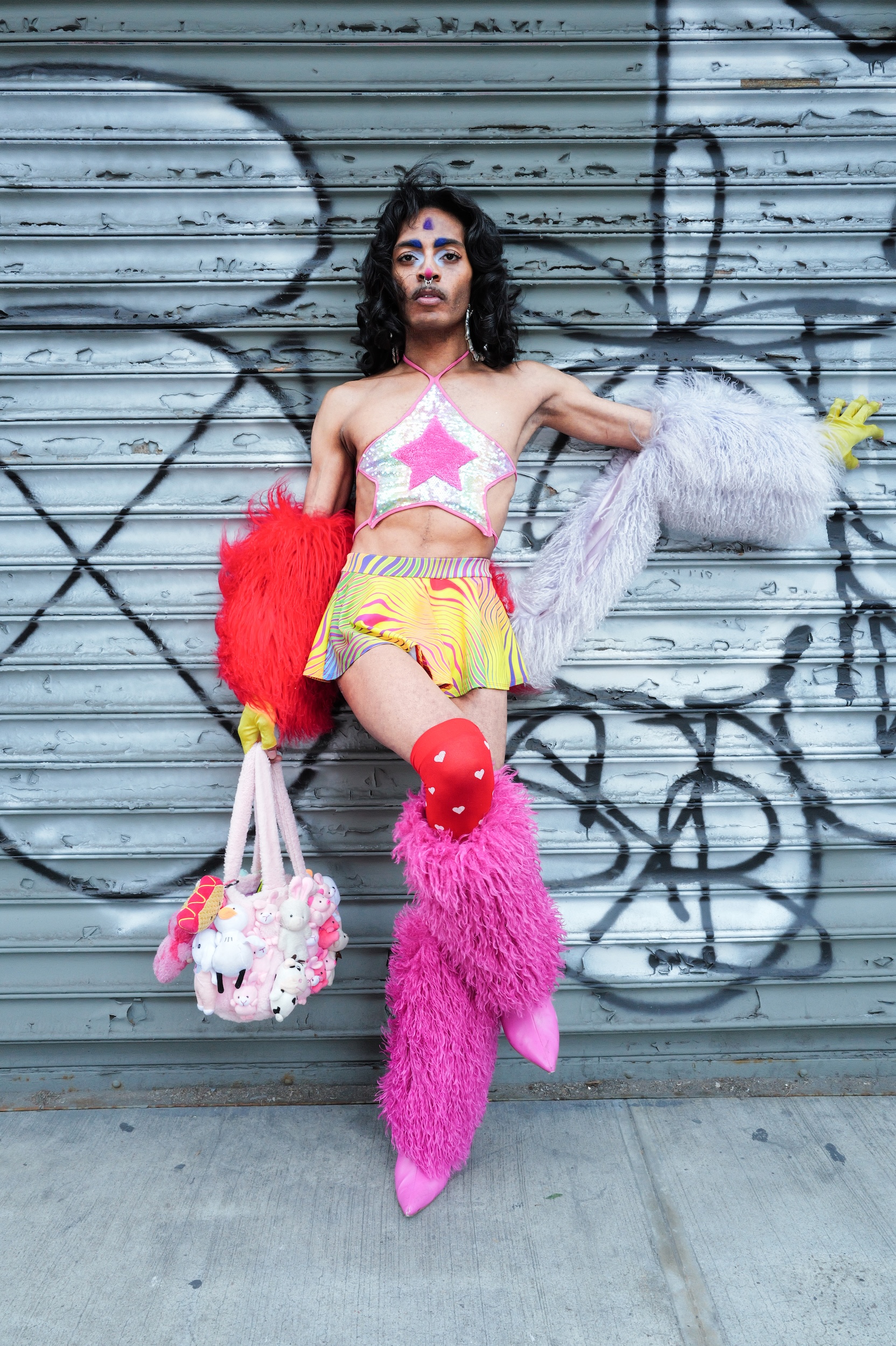 Person in colourful carnival costume with feathered fans and furry pink leg warmers against grey graffitied shutters.