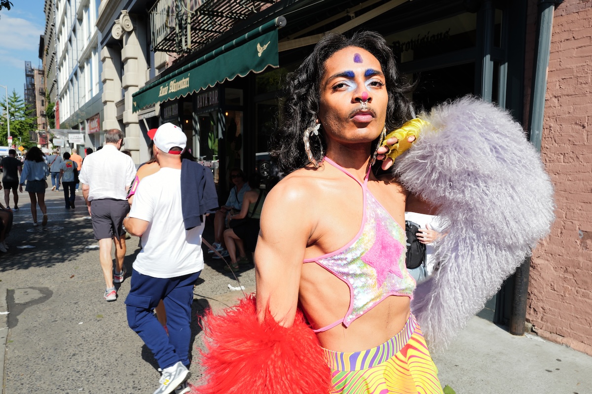 Person in colourful tie-dye outfit with white feathered wings holds red tulle on busy street with brick buildings and pedestrians.