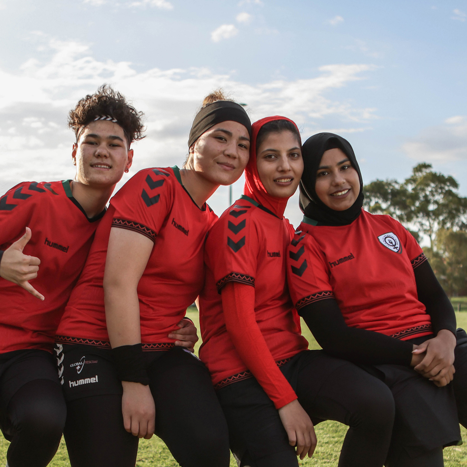 Four women in red Hummel football shirts with black chevrons sitting together on grass with blue sky and clouds behind them.