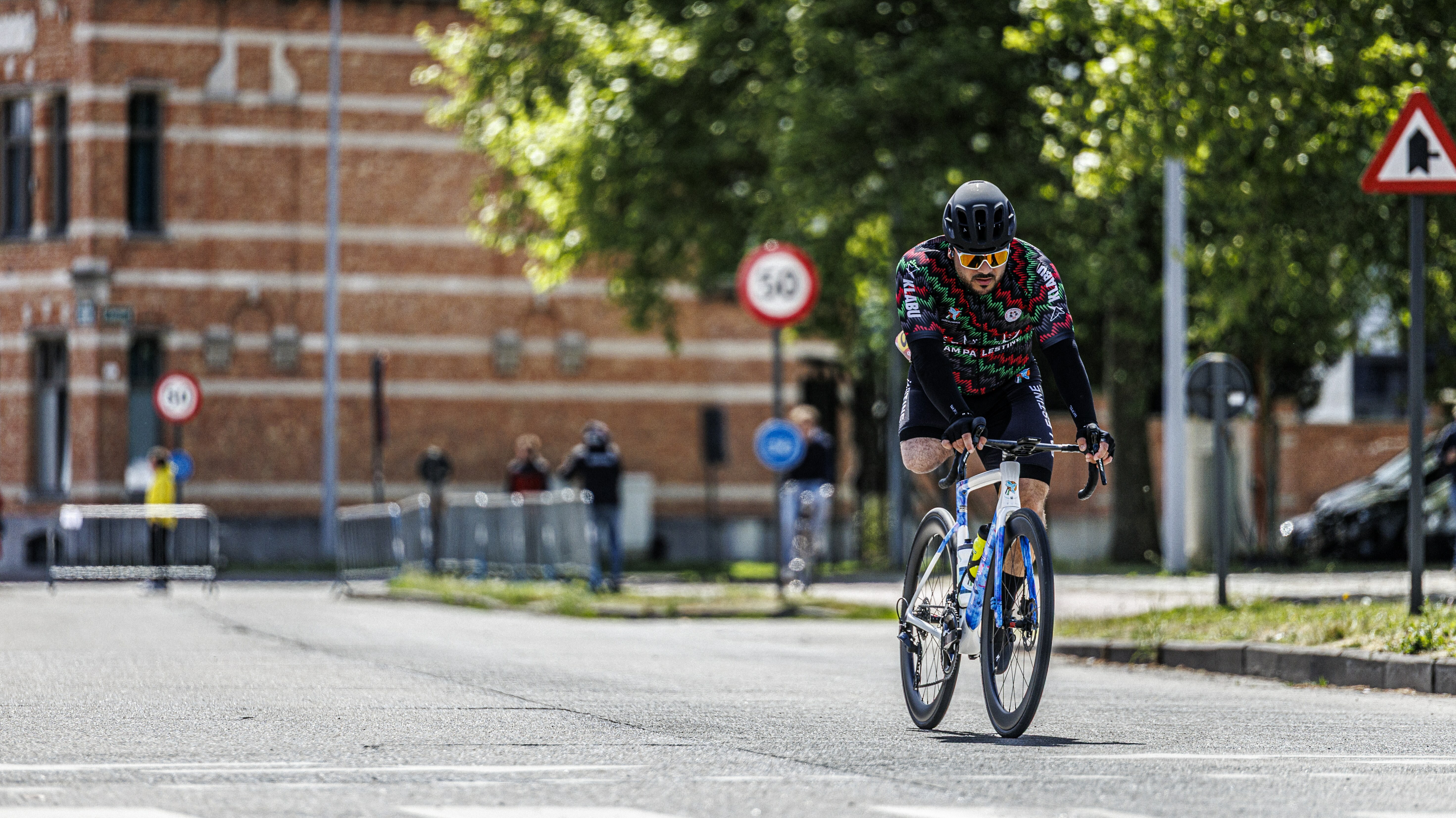 Cyclist in vibrant coloured cycling attire riding on a city street with buildings and traffic signs visible in the background.