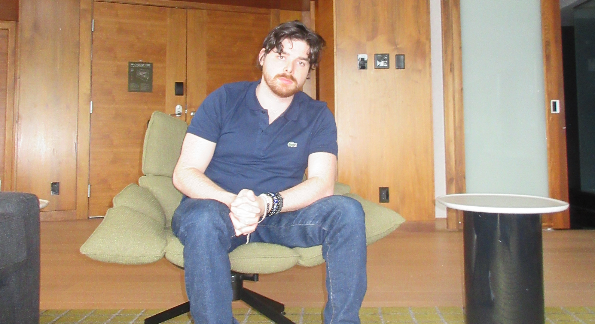 Man with glasses and beard sitting in green chair, wearing dark blue shirt and jeans in office or waiting room with wood panelling.