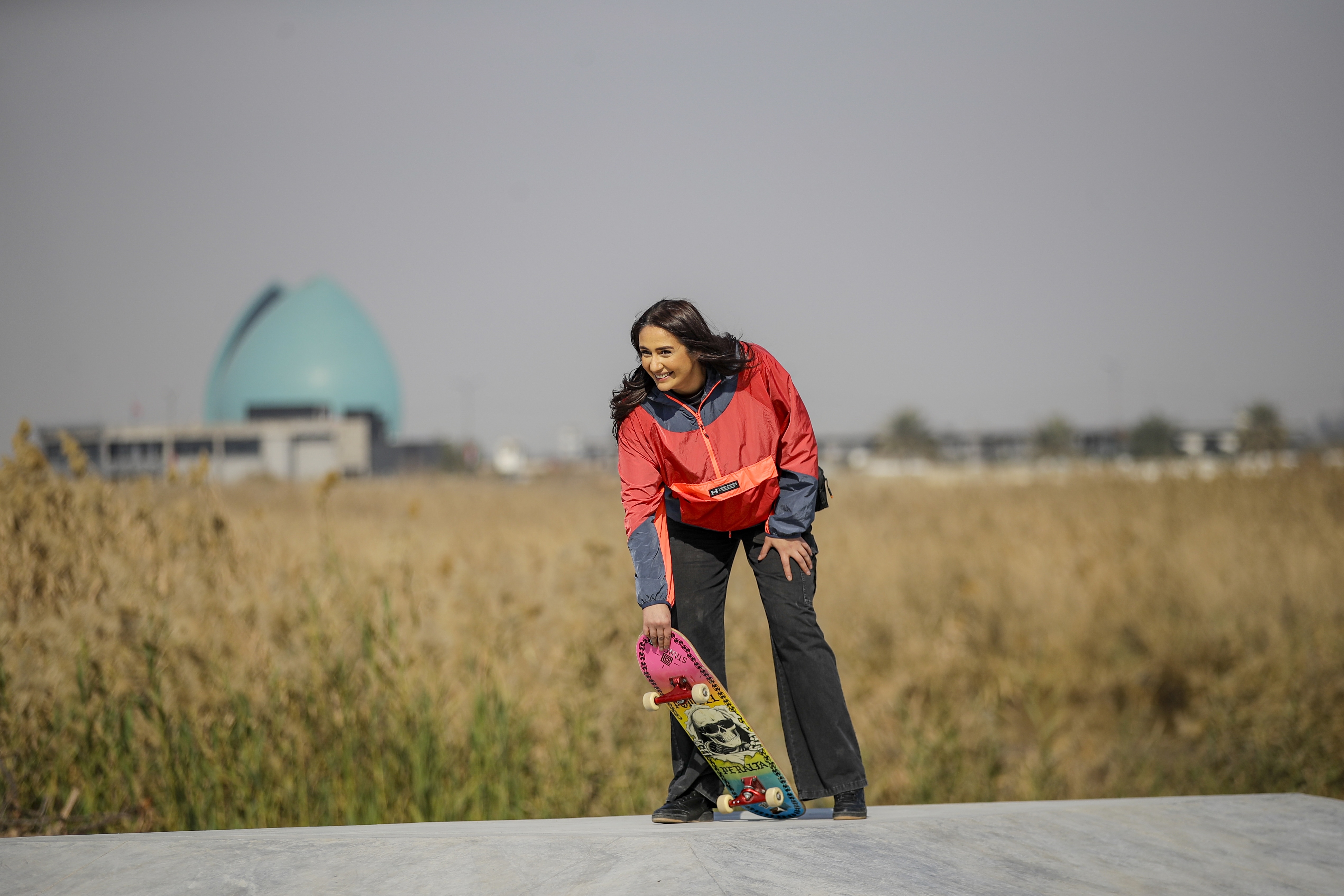 A woman in a red jacket and black trousers standing on a path in a grassy field with a domed building in the background.