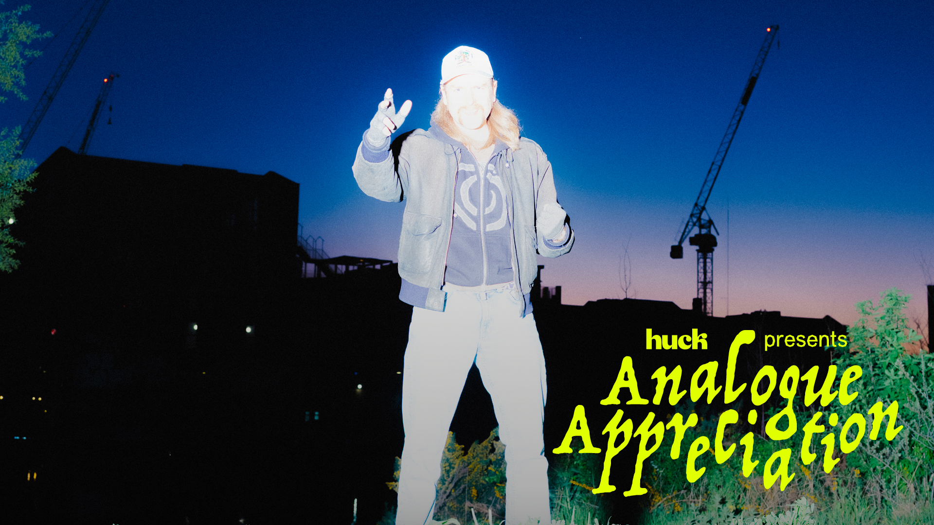White silhouette figure making peace sign against deep blue twilight sky with construction crane, yellow text reads "huck presents Analogue Appreciation"