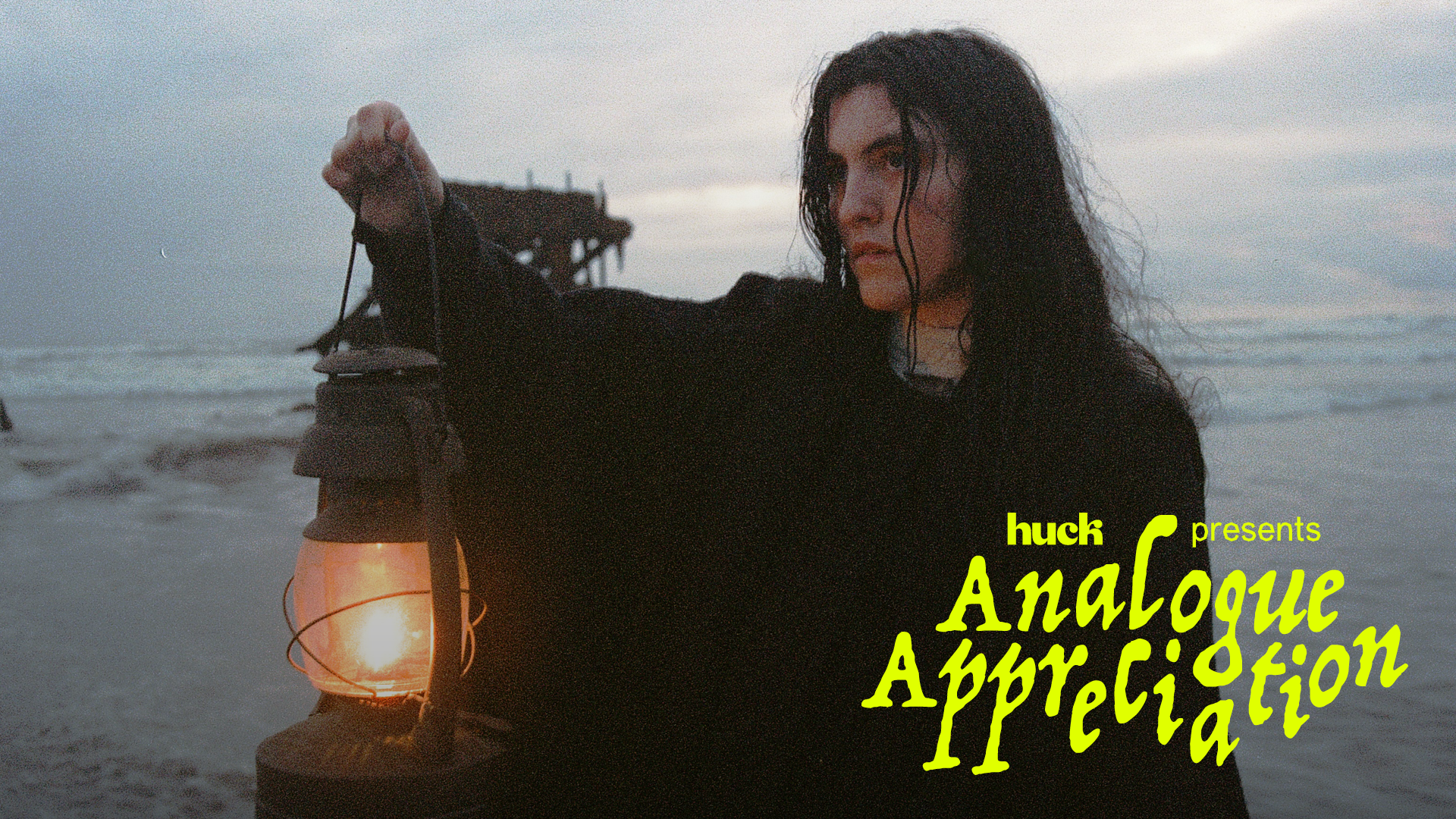 Woman with dark hair holding lit lantern on beach at dusk, shipwreck visible in background, yellow text overlay reads "Analogue Appreciation"