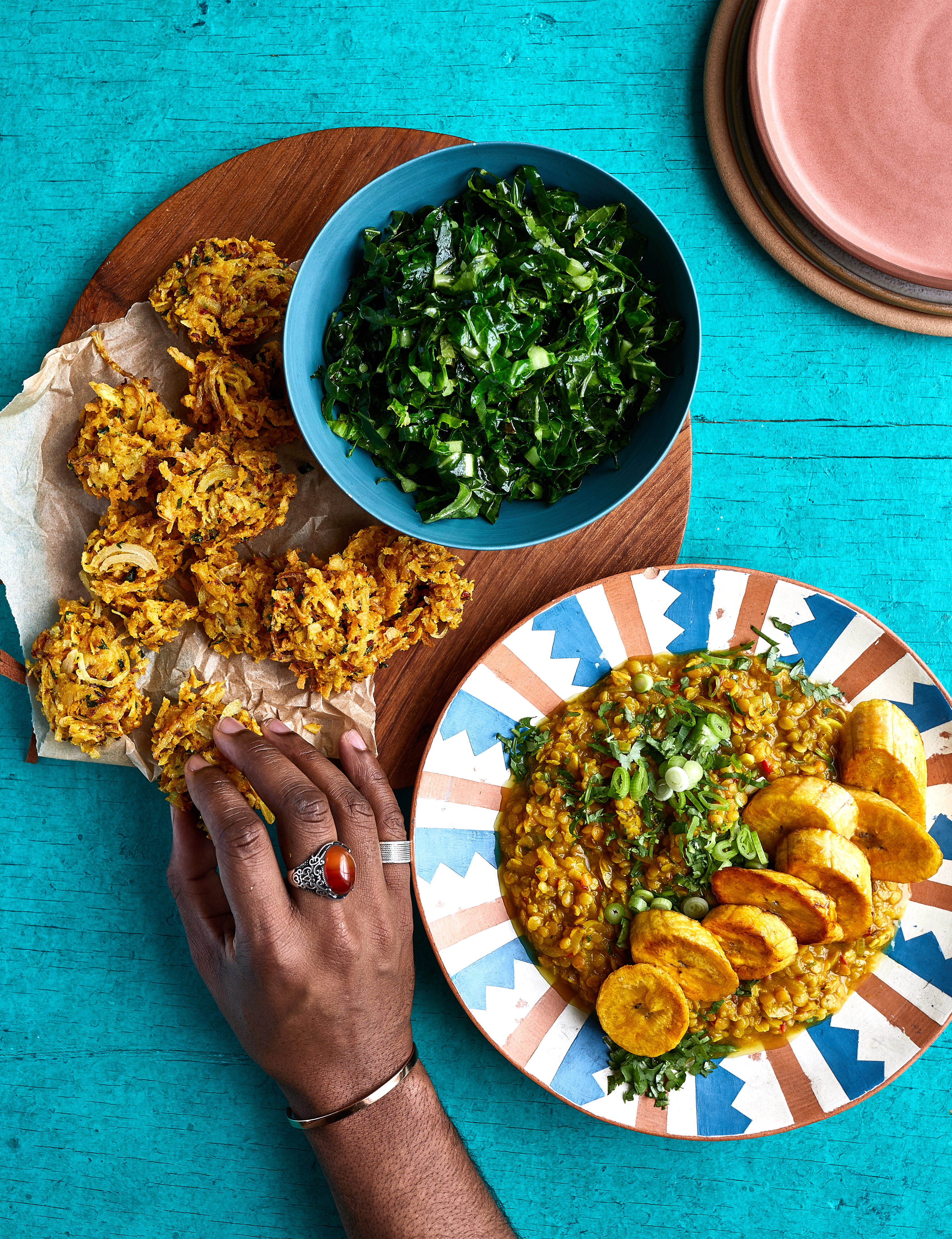 Overhead view of Caribbean meal on turquoise table: fried chicken on wooden board, green salad in blue bowl, rice dish with plantains on striped plate.