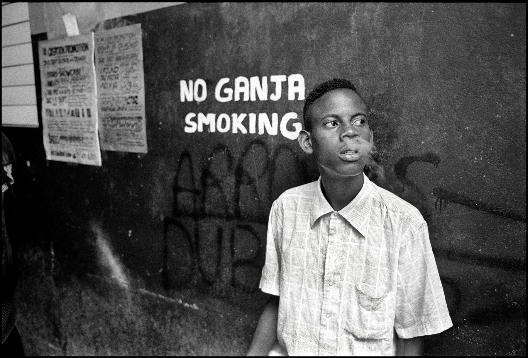 Black and white image of young man in light shirt against wall with "NO GANJA SMOKING" text and posted notices visible.