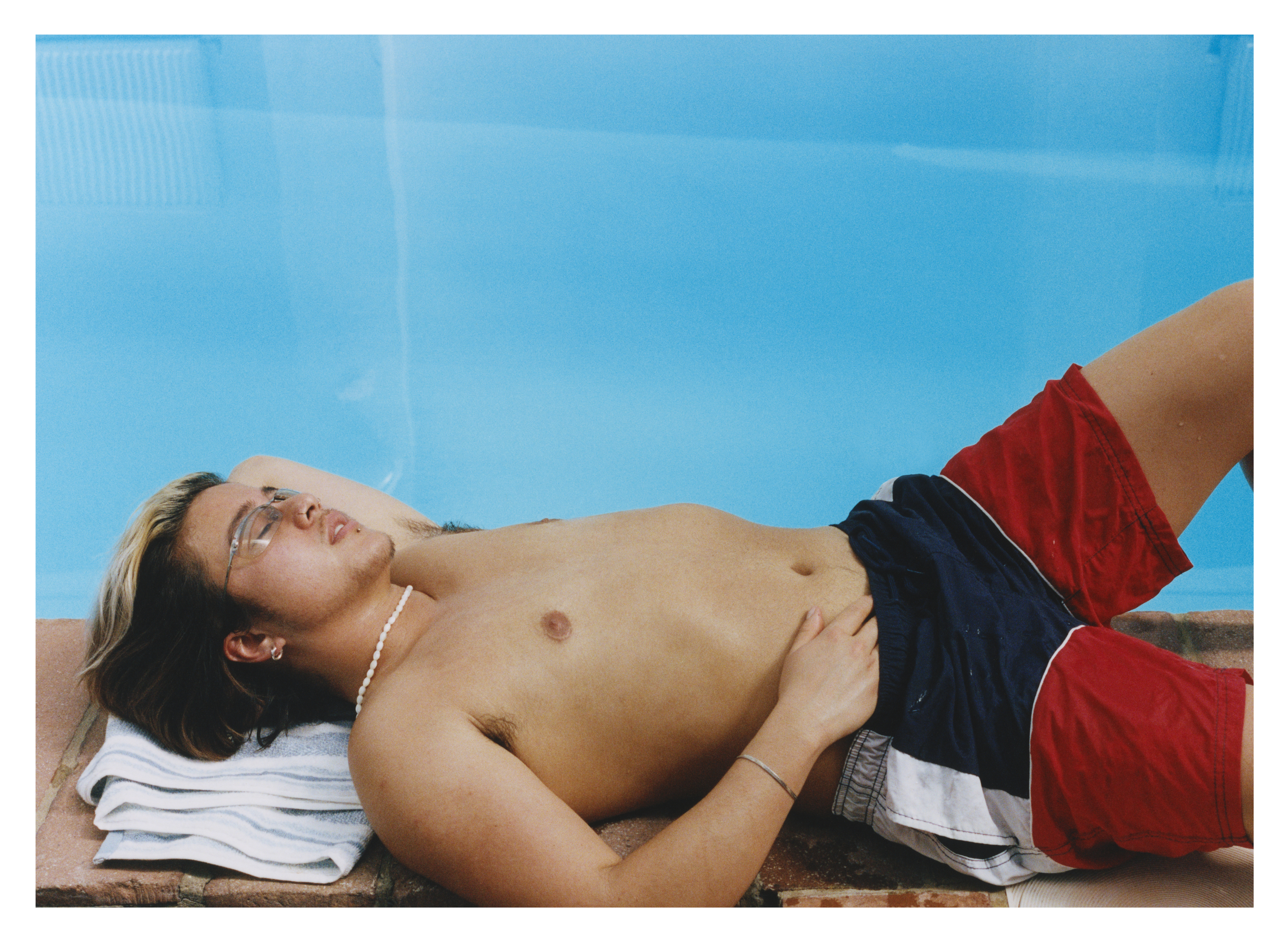 Shirtless man lying on white towel by bright blue swimming pool, wearing red and dark swim shorts, eyes closed, relaxing poolside.