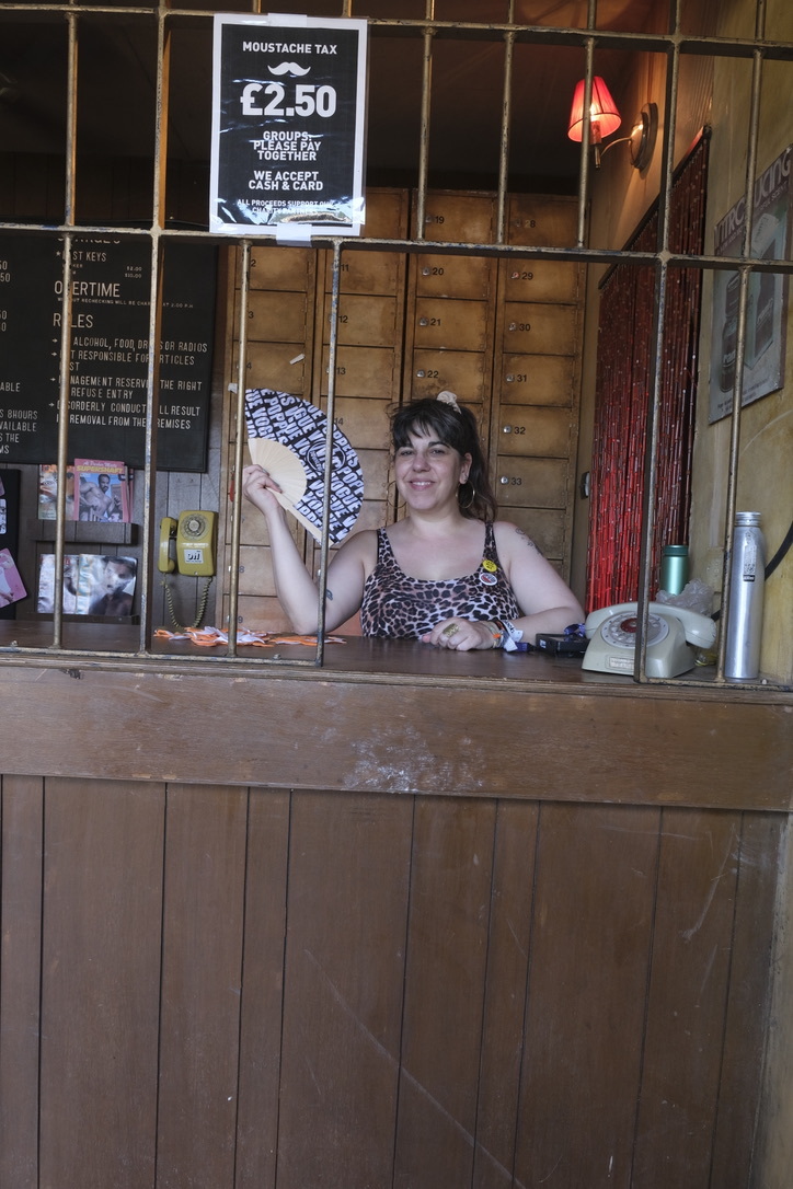 Woman behind wooden bar counter holding patterned fabric, with "Moustache Tax £2.50" sign above and shelving with bottles behind.