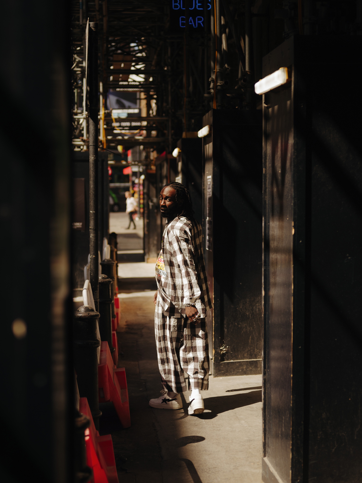 Man in chequered shirt walking through narrow alley with shops, blue neon sign visible, sunlight streaming through creating shadows.