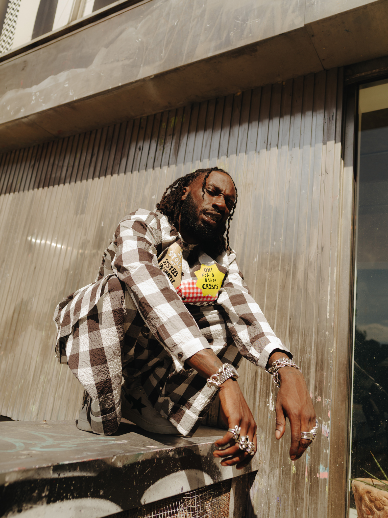 Man with dreadlocks wearing brown and white chequered shirt and jewellery, sitting on wooden steps outside building.