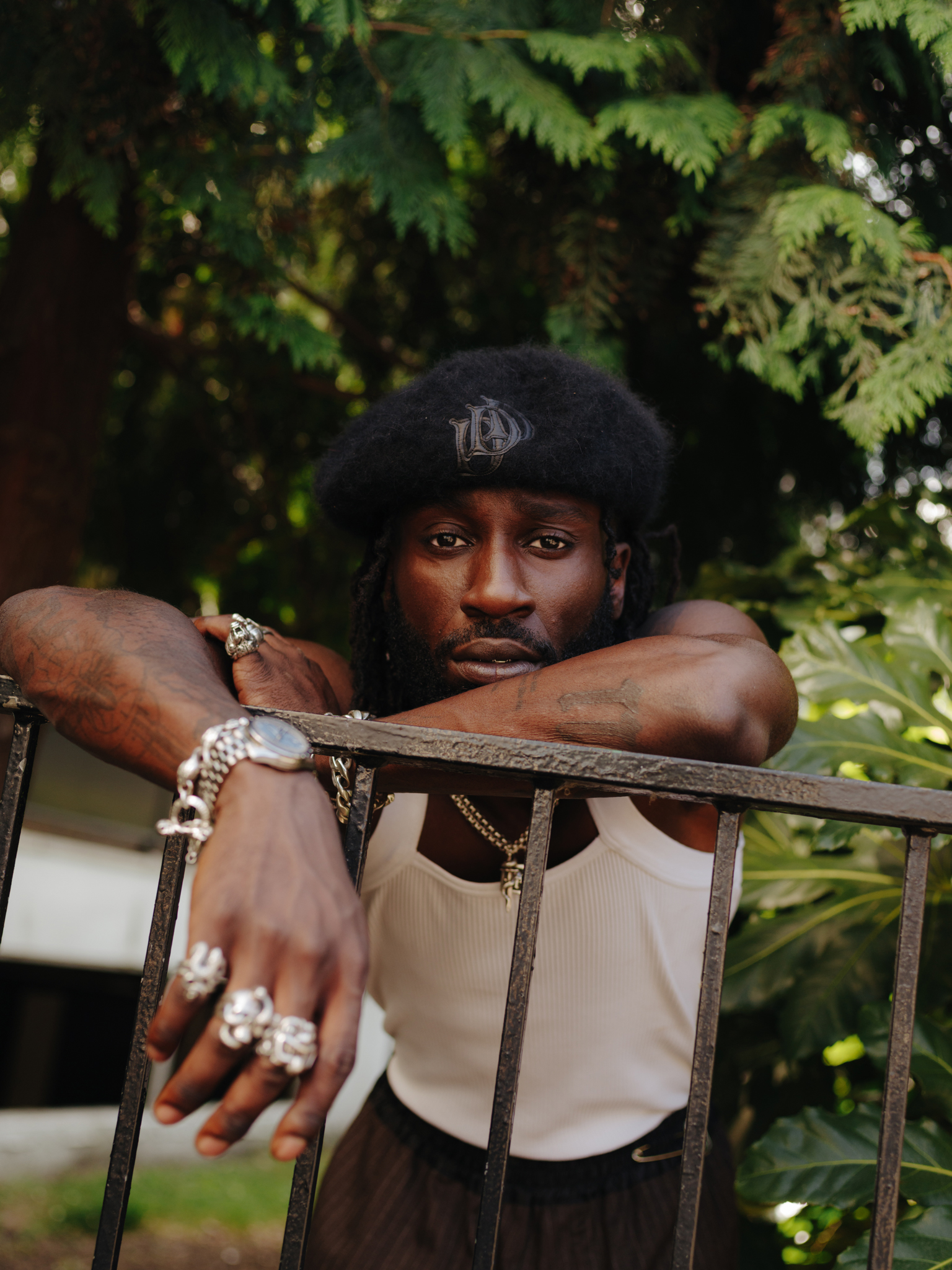 Man in black beanie and white vest leaning on metal railing, wearing rings and chains, surrounded by green foliage.