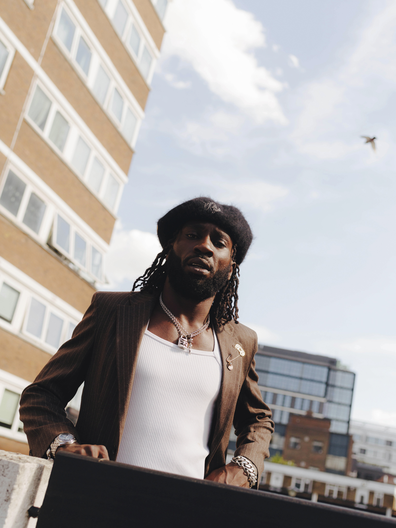 Man with dreadlocks wearing black beret, white vest, and dark jacket stands against urban backdrop of beige and grey buildings under cloudy sky.
