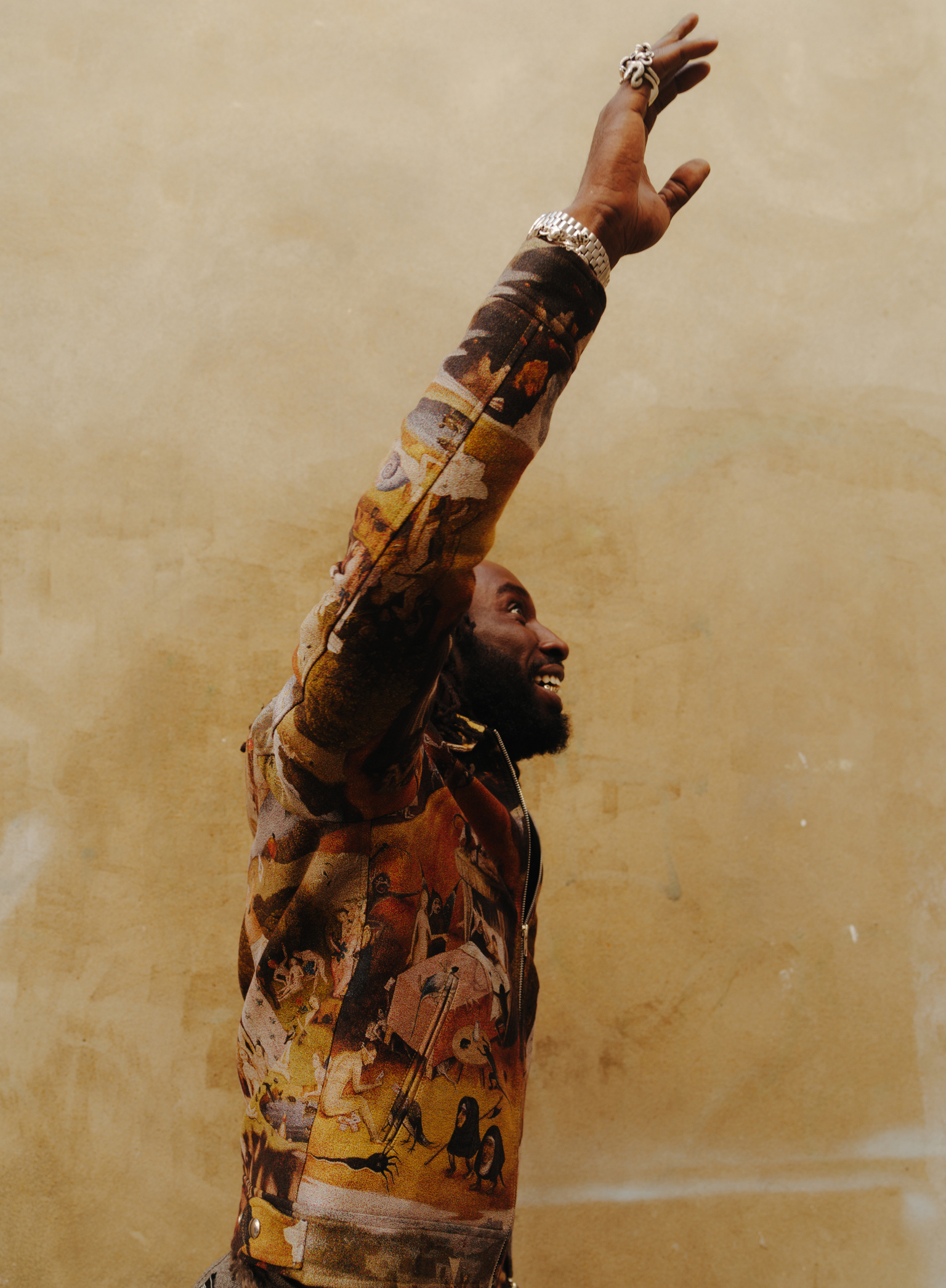 Woman with arms raised upward, wearing colourful patterned clothing and jewellery, against beige background with warm lighting.