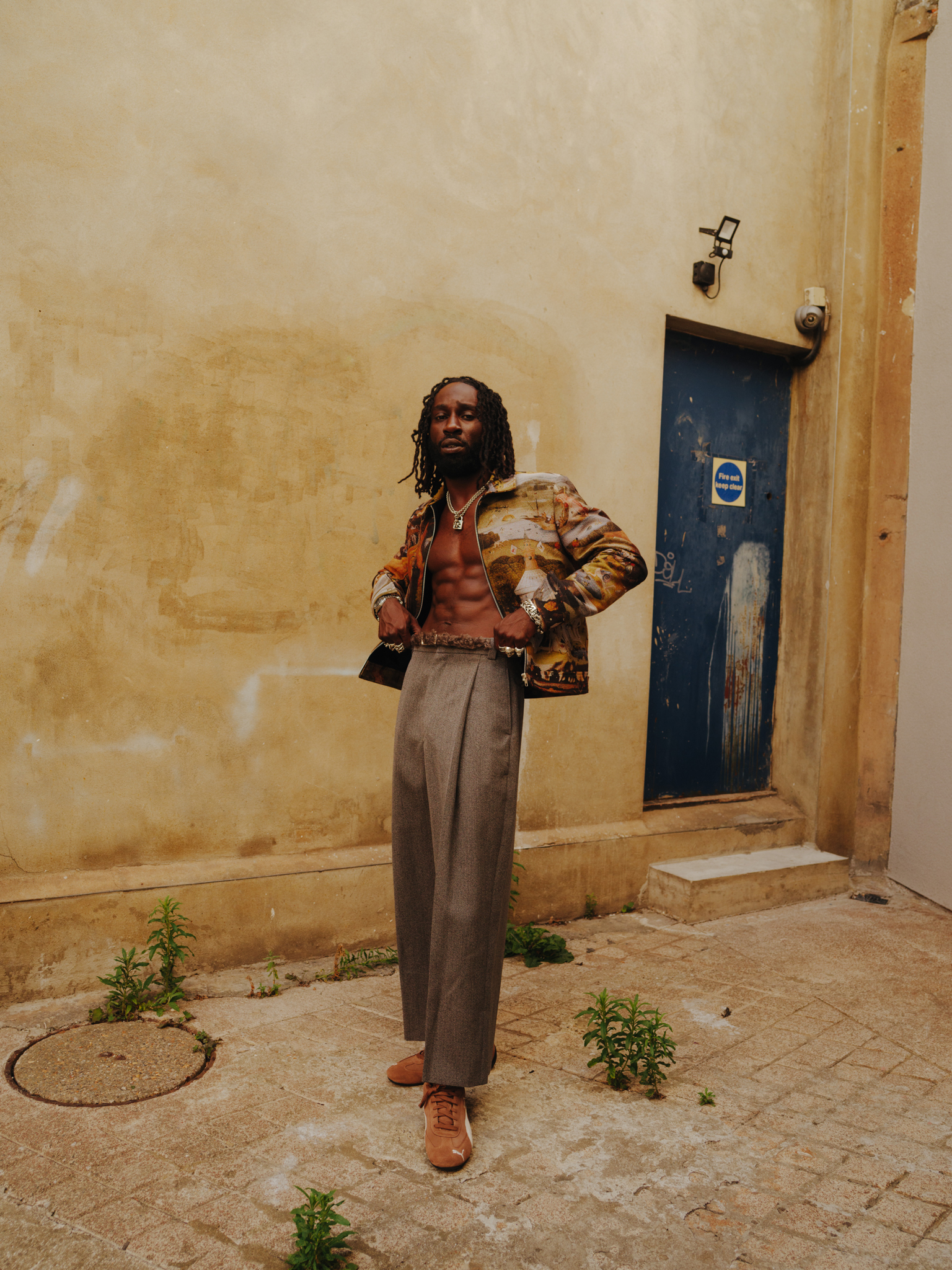 Man with dreadlocks wearing patterned jacket and wide-leg trousers stands against cream wall beside blue door with small plants nearby.