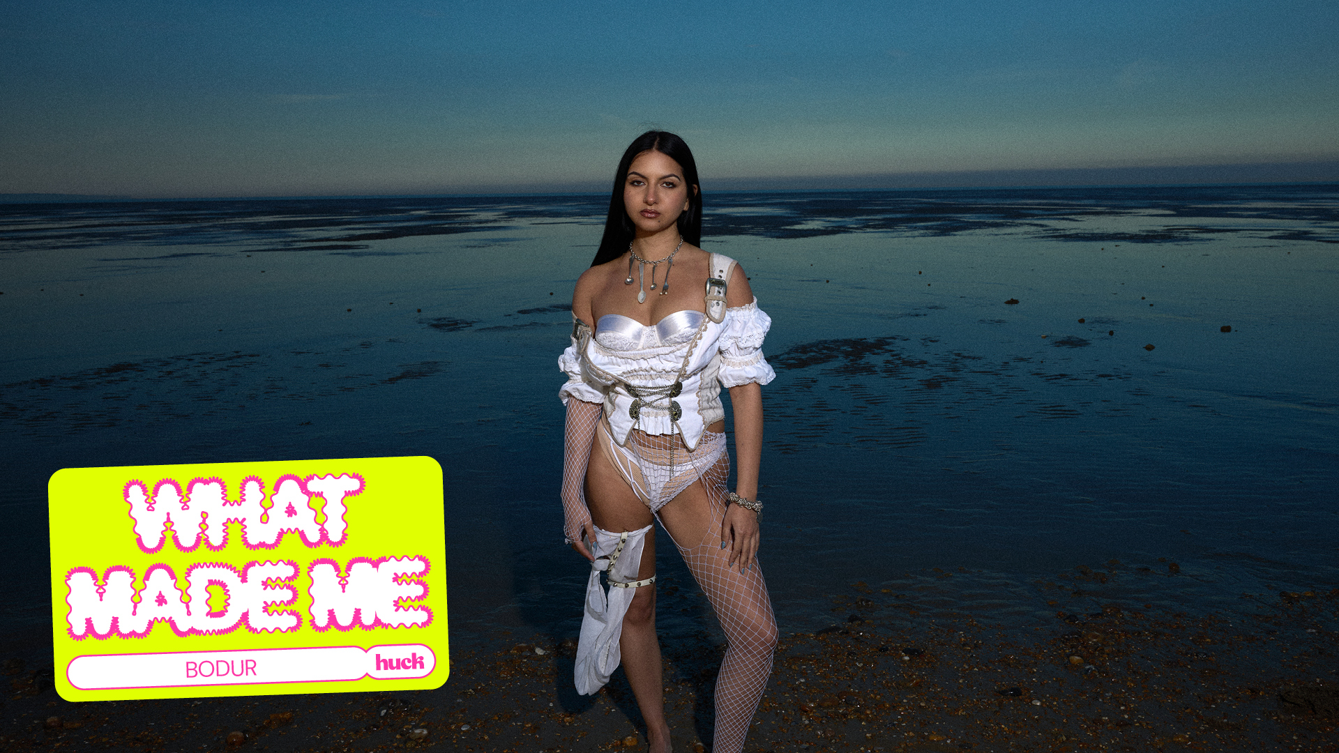 A young woman in a white blouse and shorts standing on a beach with a sign that says "What made me" in large text.