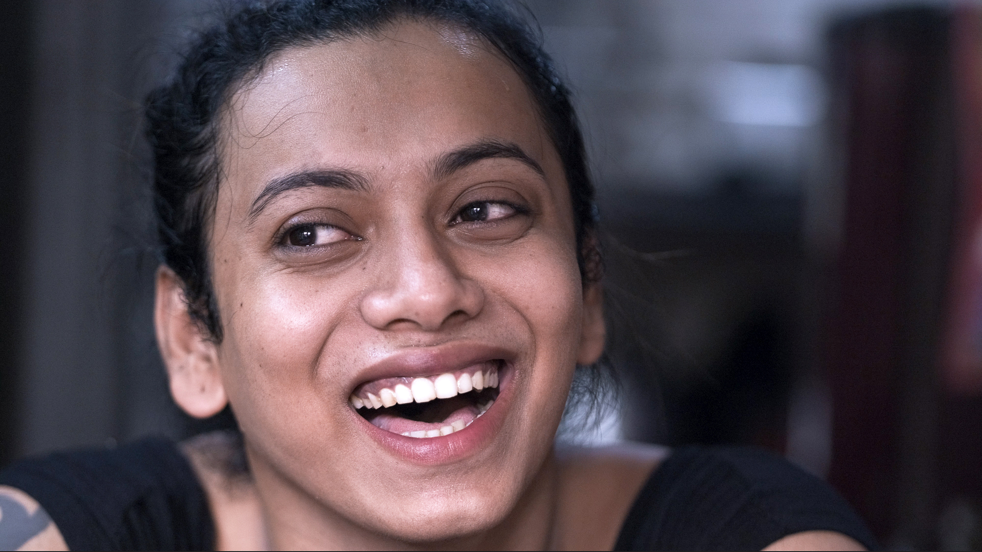 Woman with dark hair tied back, laughing with mouth open showing white teeth, looking to the right against blurred background.