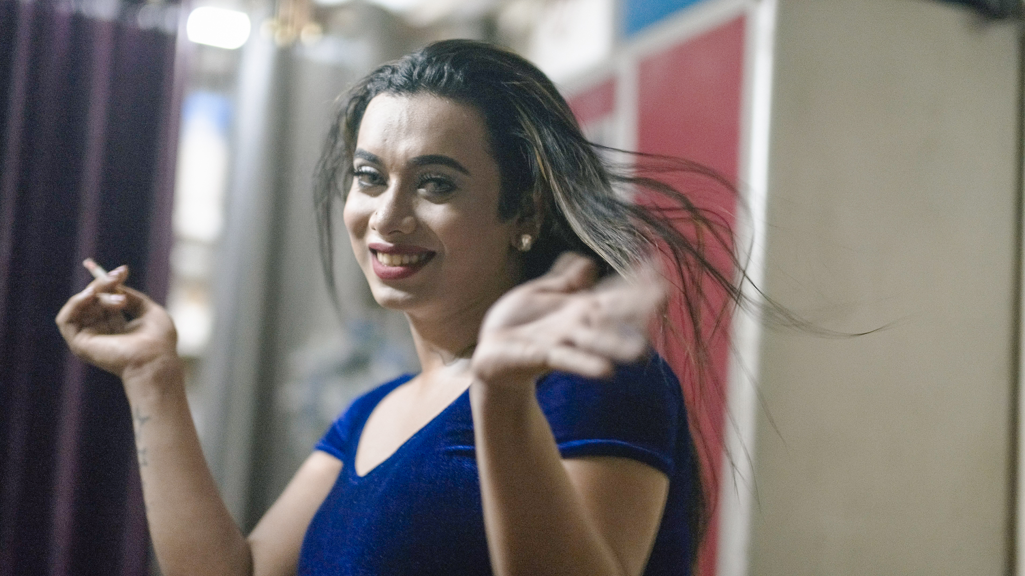 Woman with dark hair in blue top gesturing with both hands, smiling. Blurred background with colourful shapes.