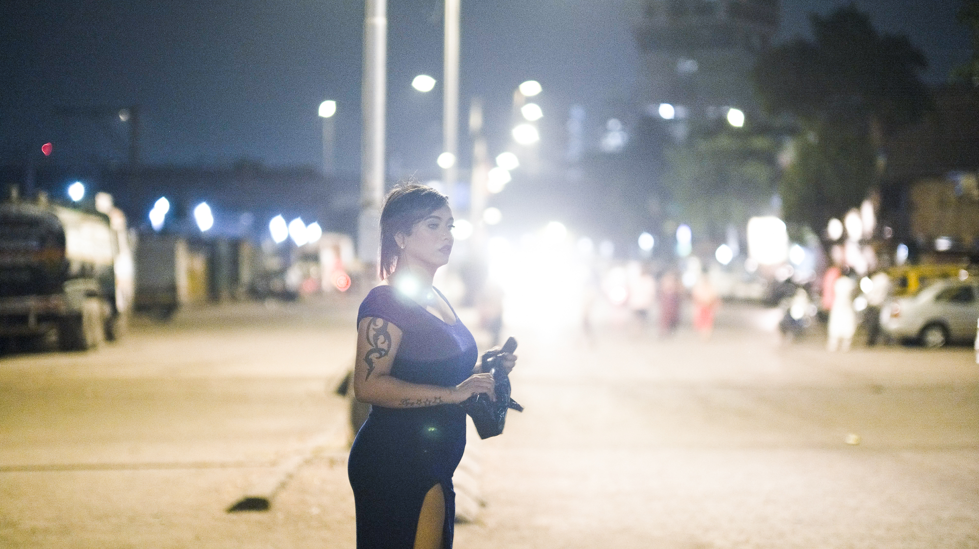 Police officer in dark uniform stands on street at night with bright lights and blurred vehicles in background.