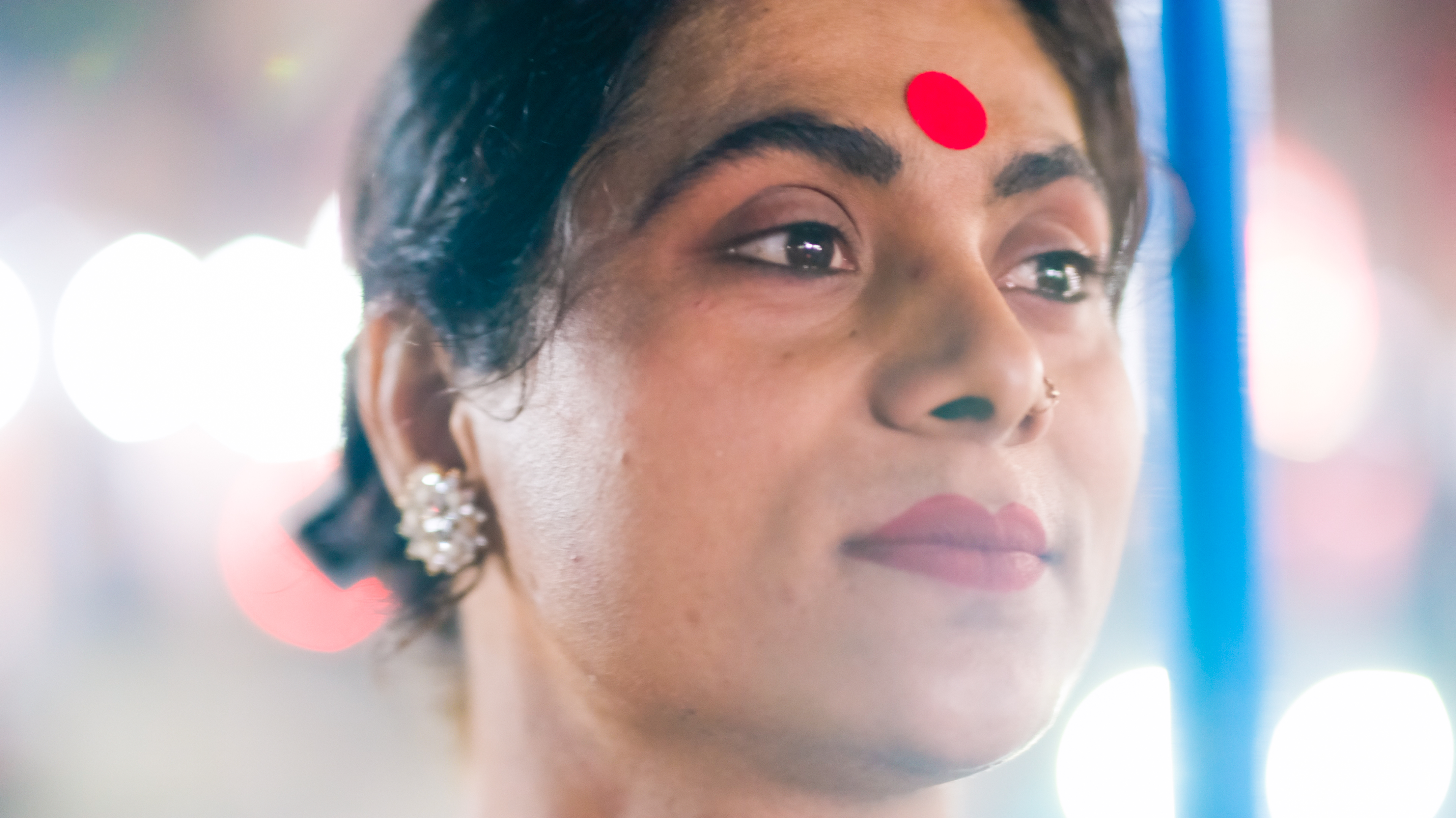 Woman with red bindi on forehead, pearl earrings, dark hair pulled back, looking upward against blurred colourful background.