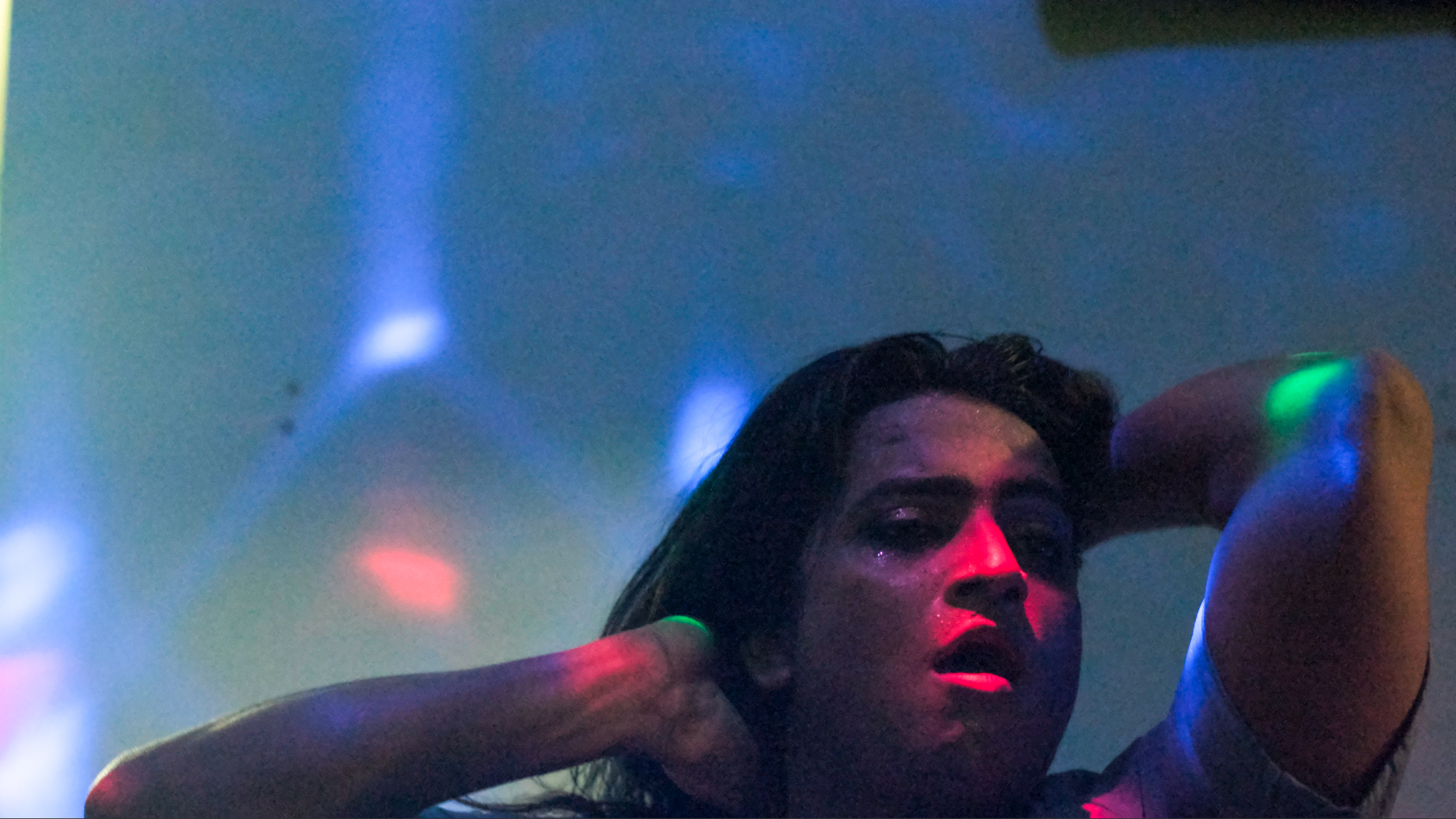 Woman with long dark hair in coloured lighting - red, blue, and green beams create dramatic shadows across her face and raised arms.