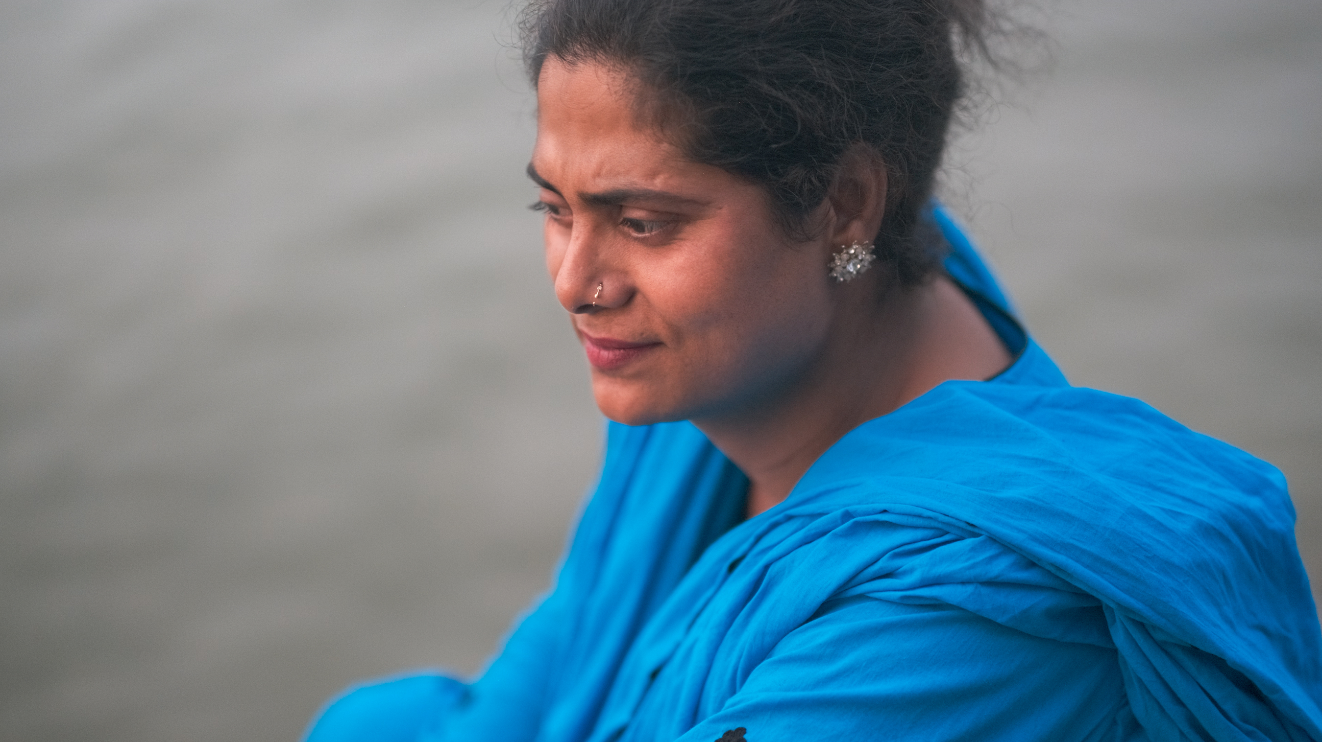 Woman in profile wearing bright blue garment, dark hair pulled back, small earrings visible, grey background.