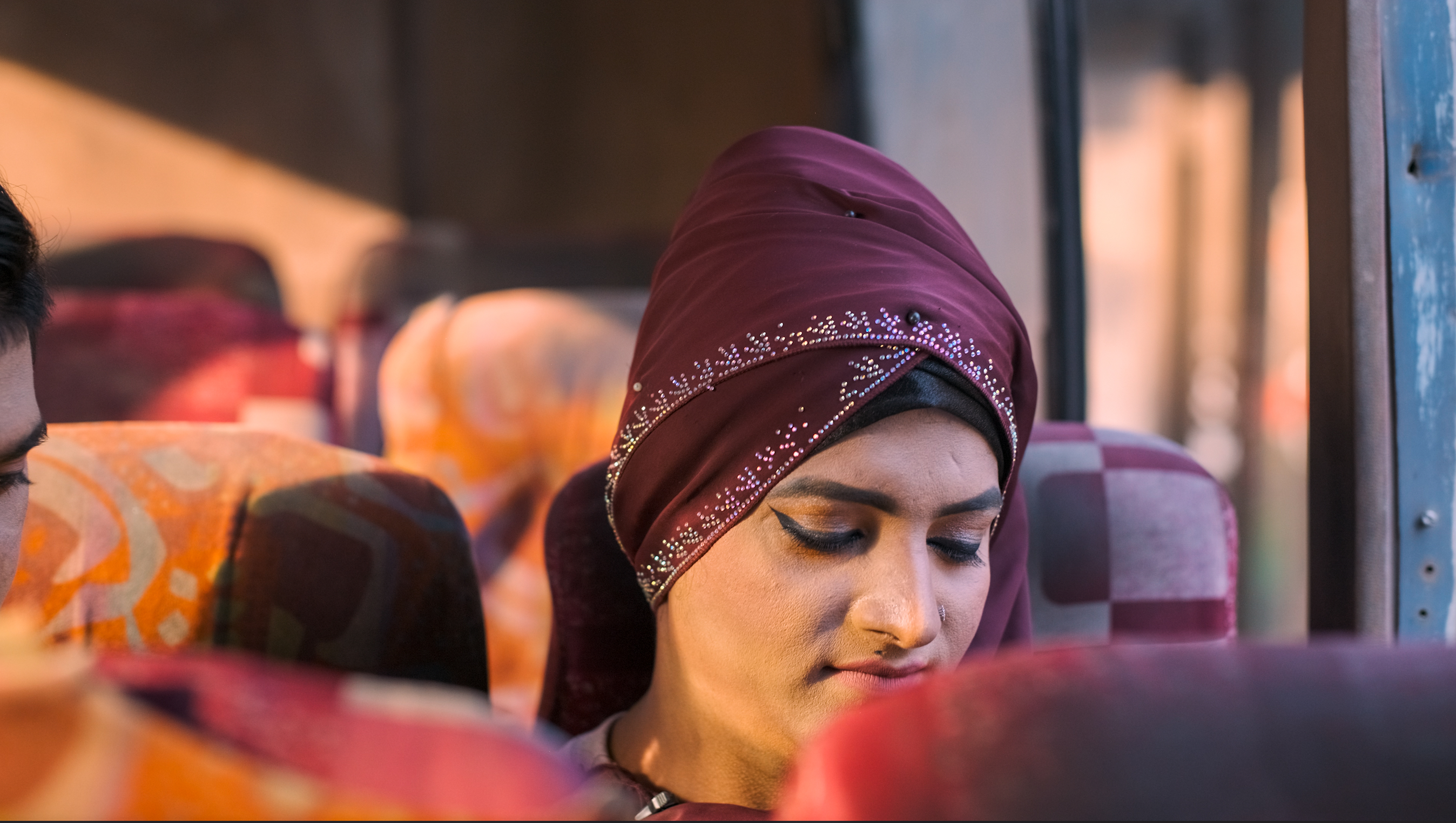 Woman in burgundy headscarf sitting on bus, eyes closed, with blurred orange and red seats visible in background.