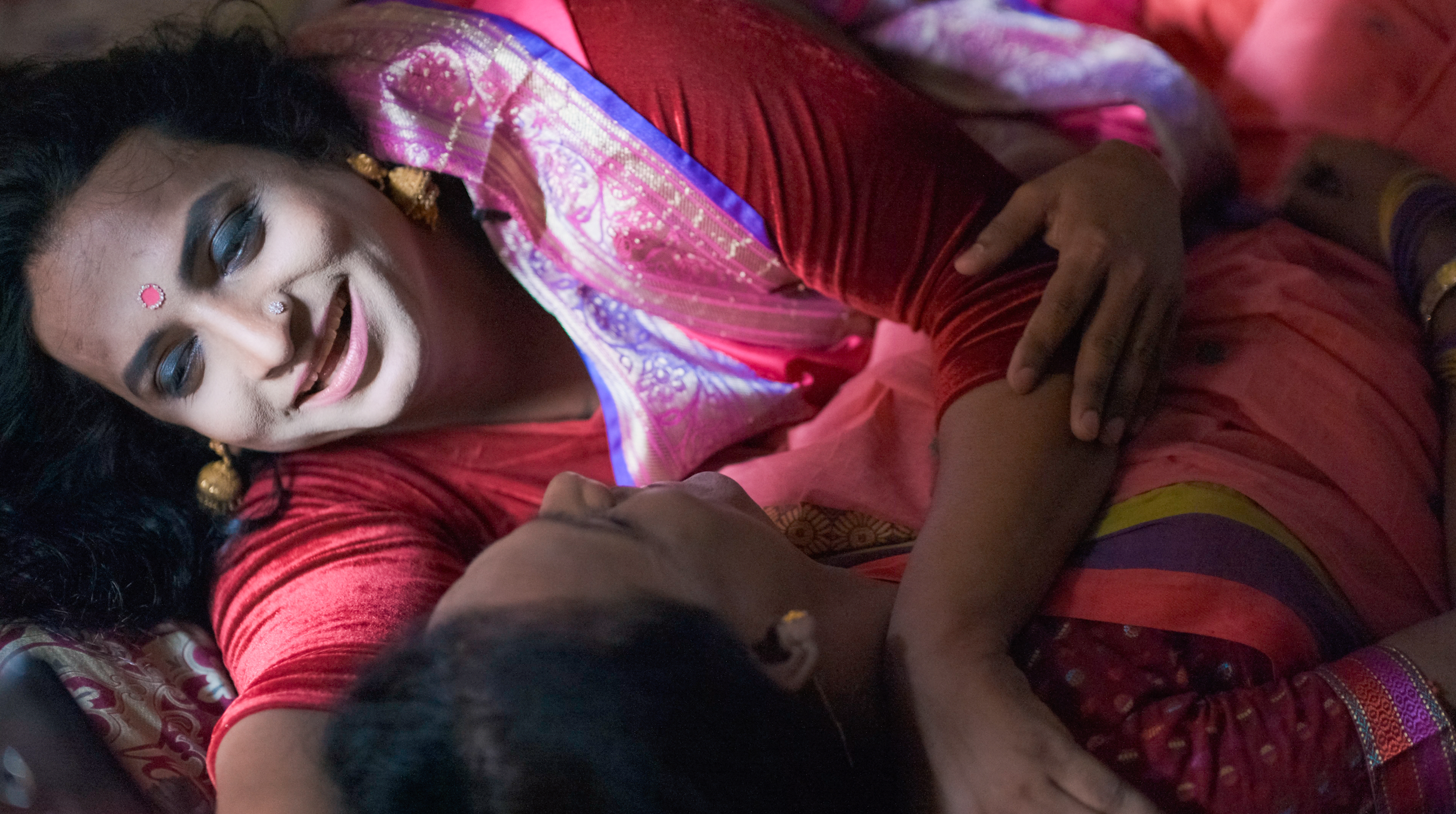 Woman in red sari lying on colourful fabric, smiling at camera with hand positioned on her torso.