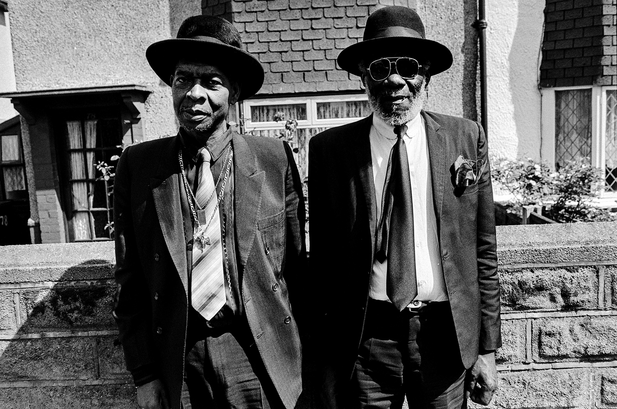 Two elderly Black men in suits and hats standing on concrete steps outside brick building, black and white photograph.