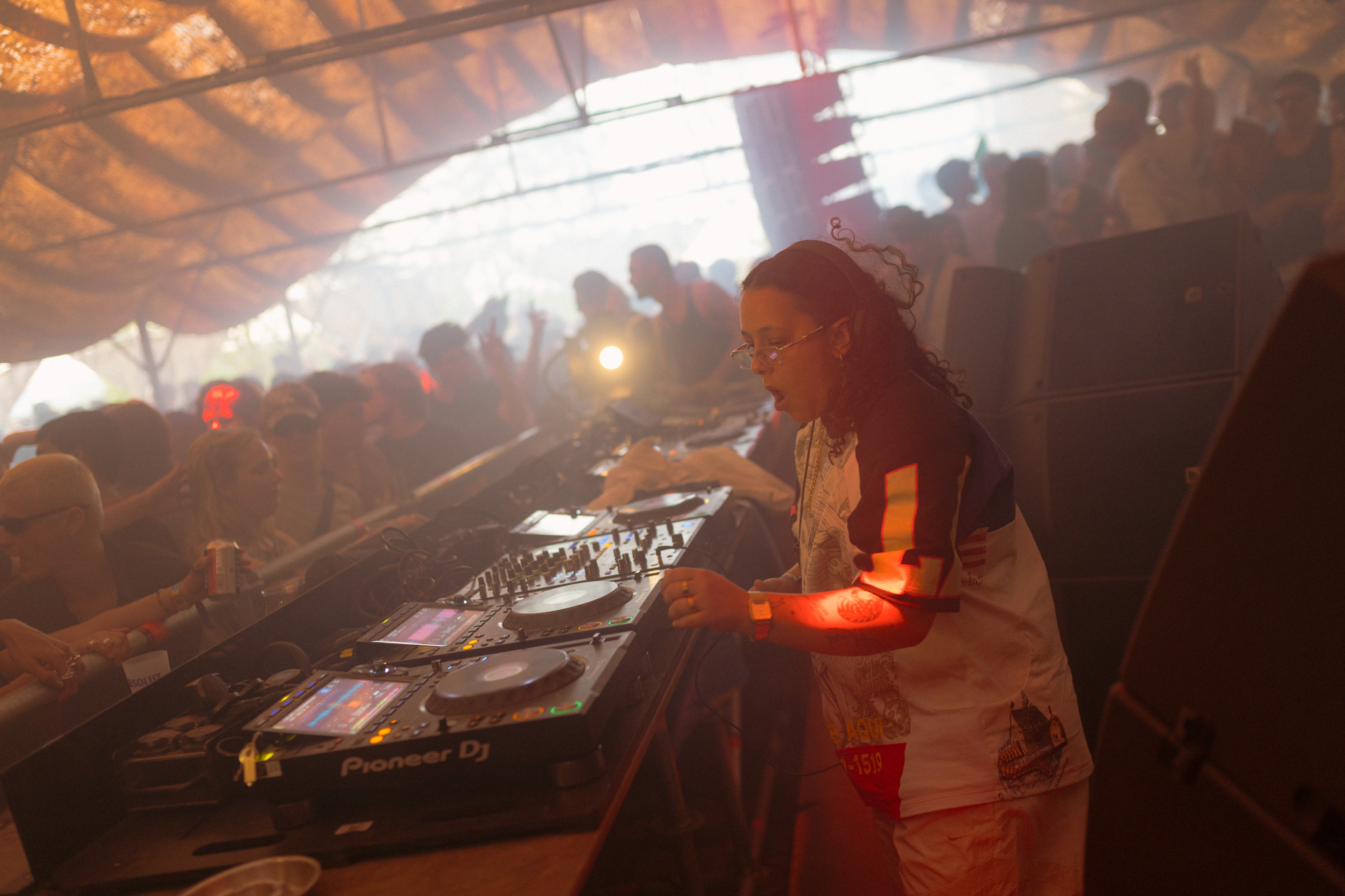 Blurred silhouette of a person behind DJ equipment, surrounded by a crowd and colourful lighting in a nightclub setting.